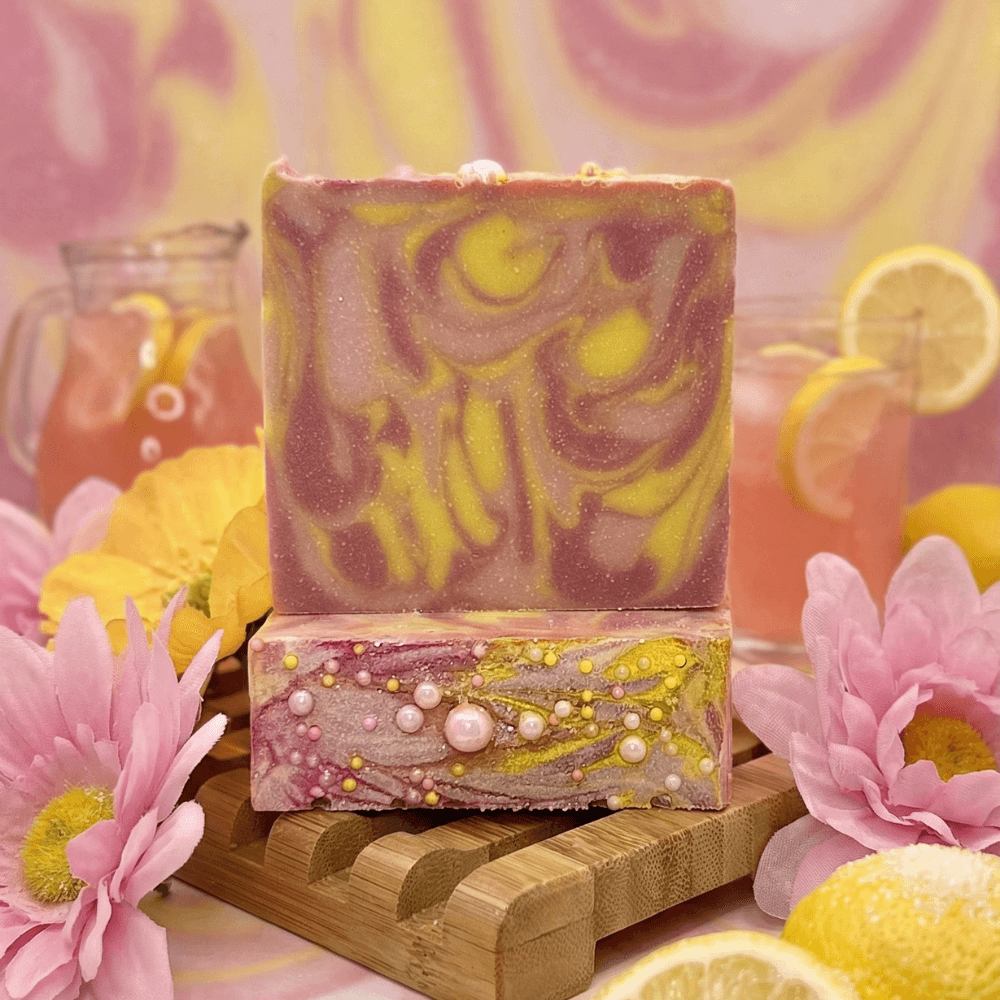Two stacked bars of swirled pink and yellow soap with marbled top and pearl-studded base, displayed on bamboo holder with pink flowers, lemon slices, and pitcher in background.