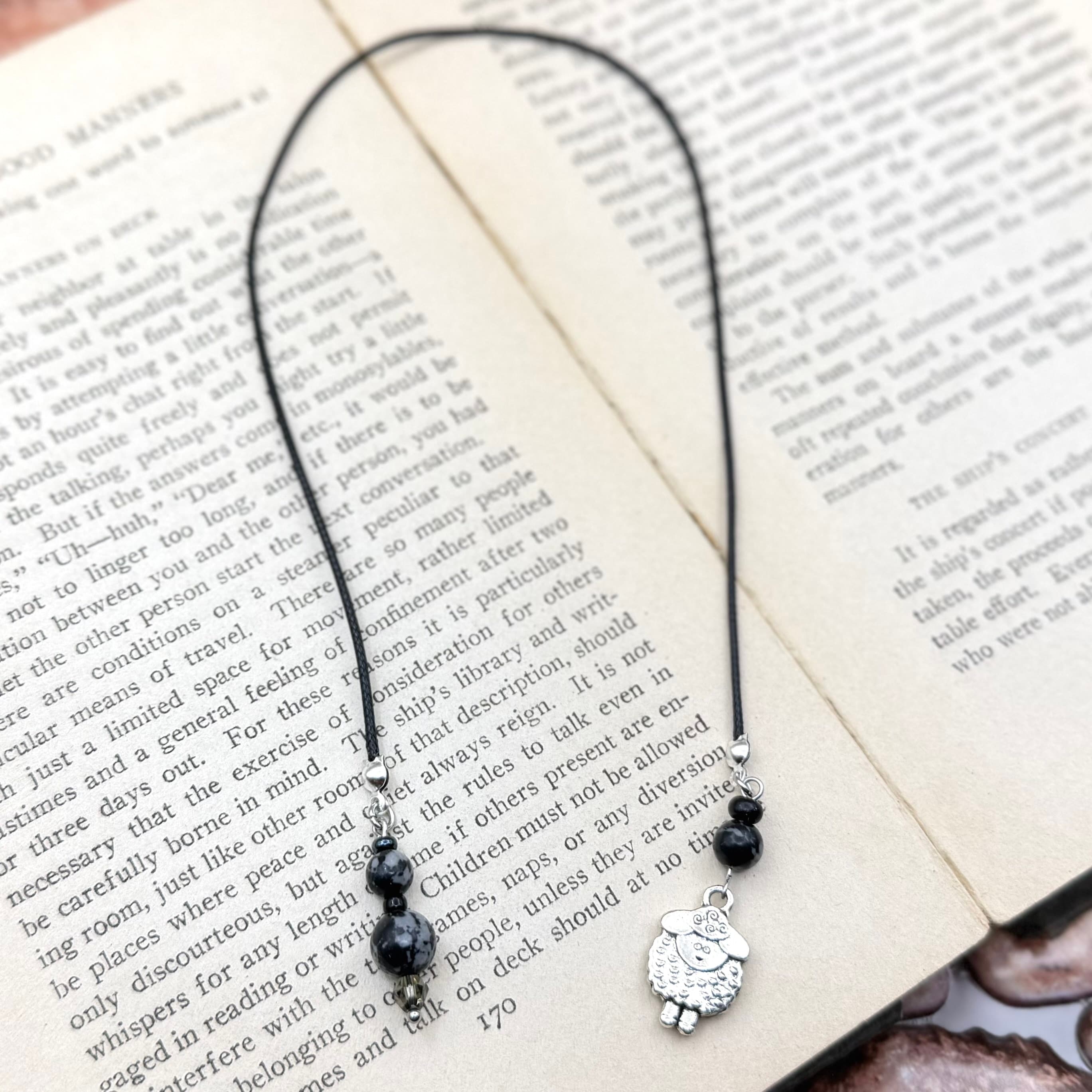 Handmade bookmark with black cord forming a heart shape over open book pages. Features black beaded accents and silver sheep charm dangling at bottom center.
