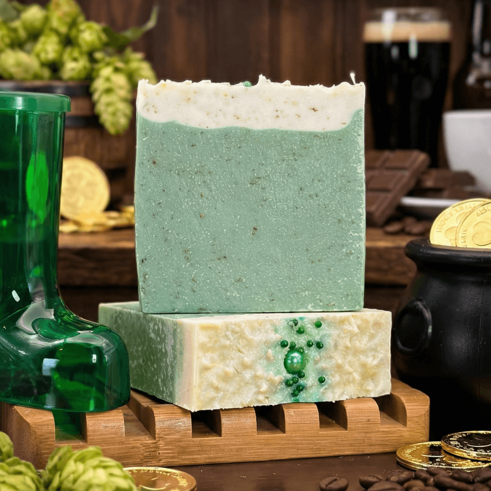 Two handcrafted soap bars stacked on a wooden soap dish. The mint-green bar has a creamy white top with specks. The cream-colored bar features green decorative beads embedded in the surface.