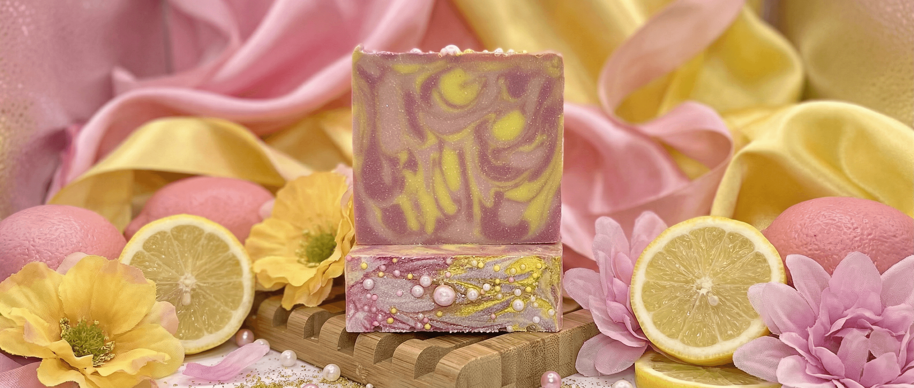Two-toned artisan soap bar with pink and yellow marbled swirl pattern, displayed on wooden stand with fresh lemon slices, pink flowers, and pearl accents in soft pastel styling.