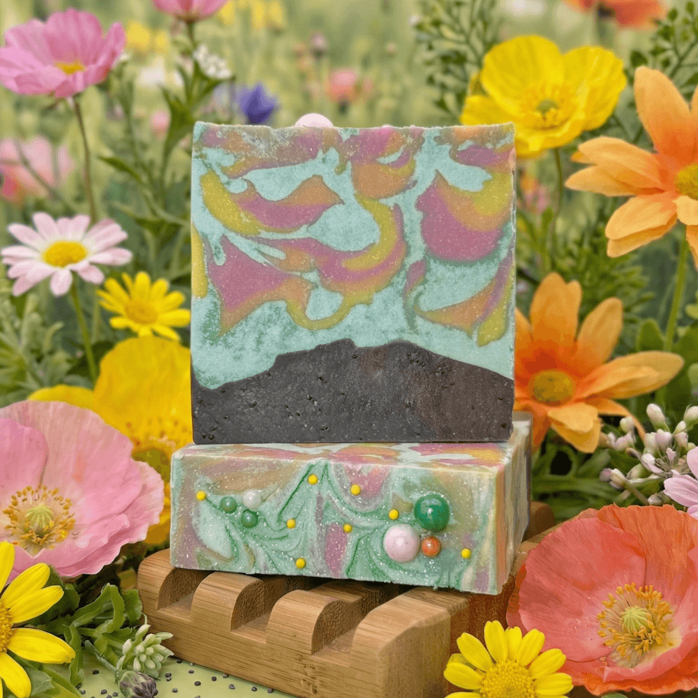 Two stacked handcrafted soap bars on wooden holder amid wildflowers. Top bar features swirled mint, pink, orange layers with dark charcoal base. Bottom bar has pastel swirls with colorful sphere embeds.