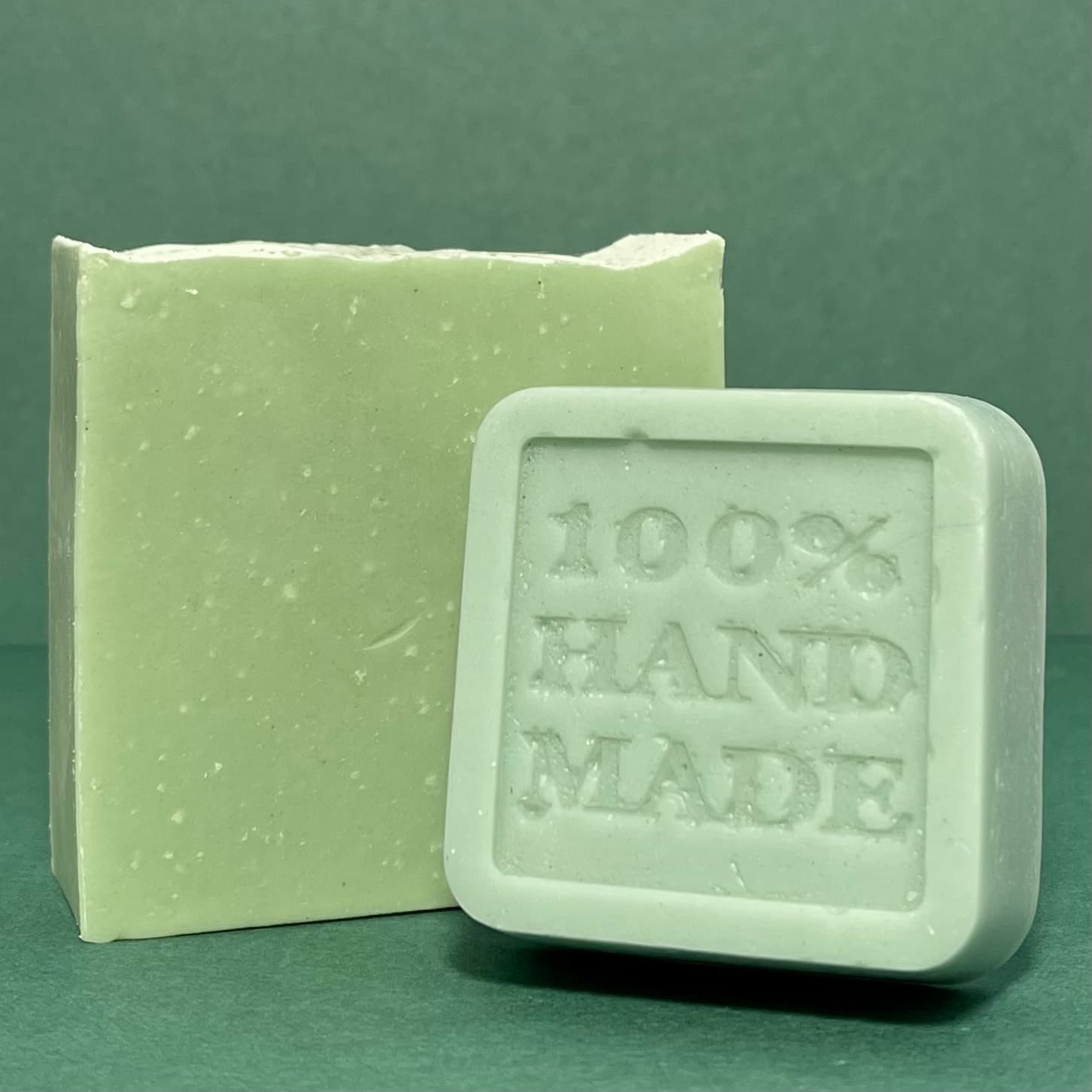 Light sage green rectangular soap bar with speckled texture next to a white square stamped with '100% HAND MADE', displayed against a teal background.