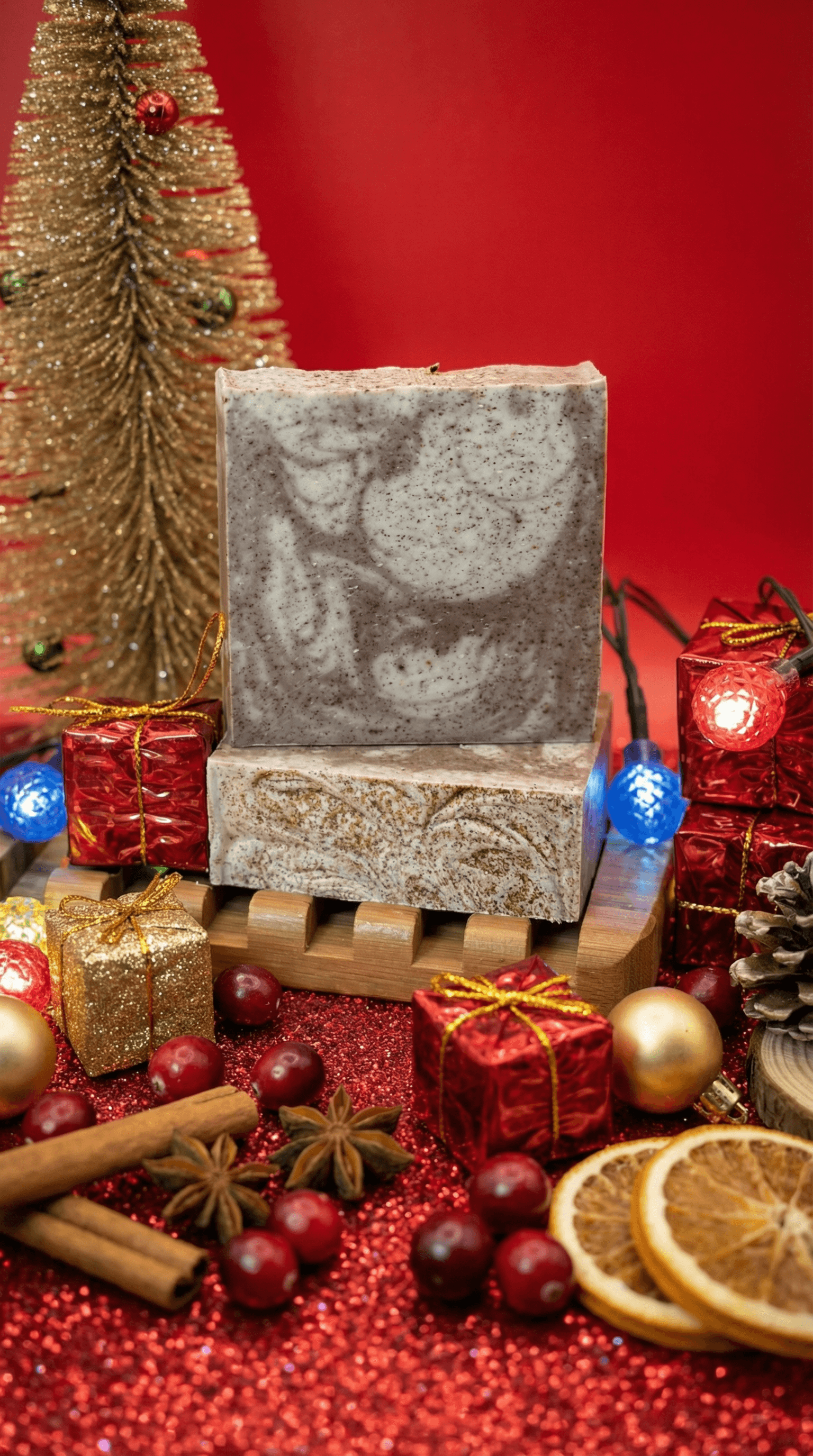 Two bars of handcrafted soap with brown speckled swirls on cream base, displayed on wooden soap dish among festive holiday decorations including miniature tree, ornaments, cinnamon sticks, dried citrus, and cranberries against red glittery background.