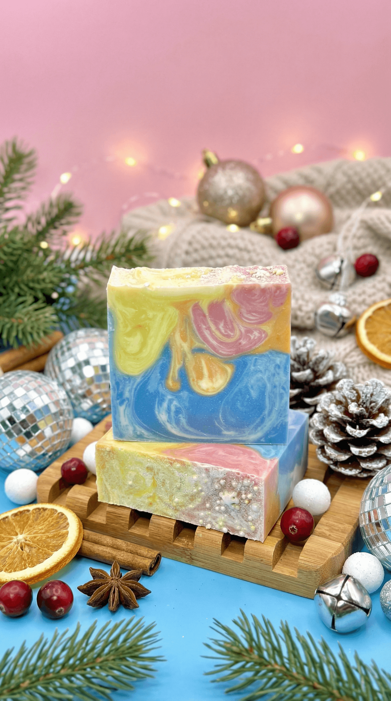 Two swirled soap bars stacked on a wooden soap dish, featuring marbled blue, yellow, pink, and cream colors. Styled with holiday decor including pine branches, dried oranges, cinnamon sticks, berries, silver ornaments, and frosted pinecones.
