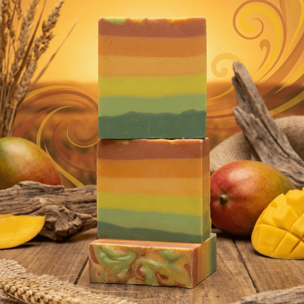 Three stacked handcrafted soap bars with horizontal gradient layers in brown, orange, yellow, and teal-green tones, displayed with fresh mango and driftwood on rustic wooden surface.