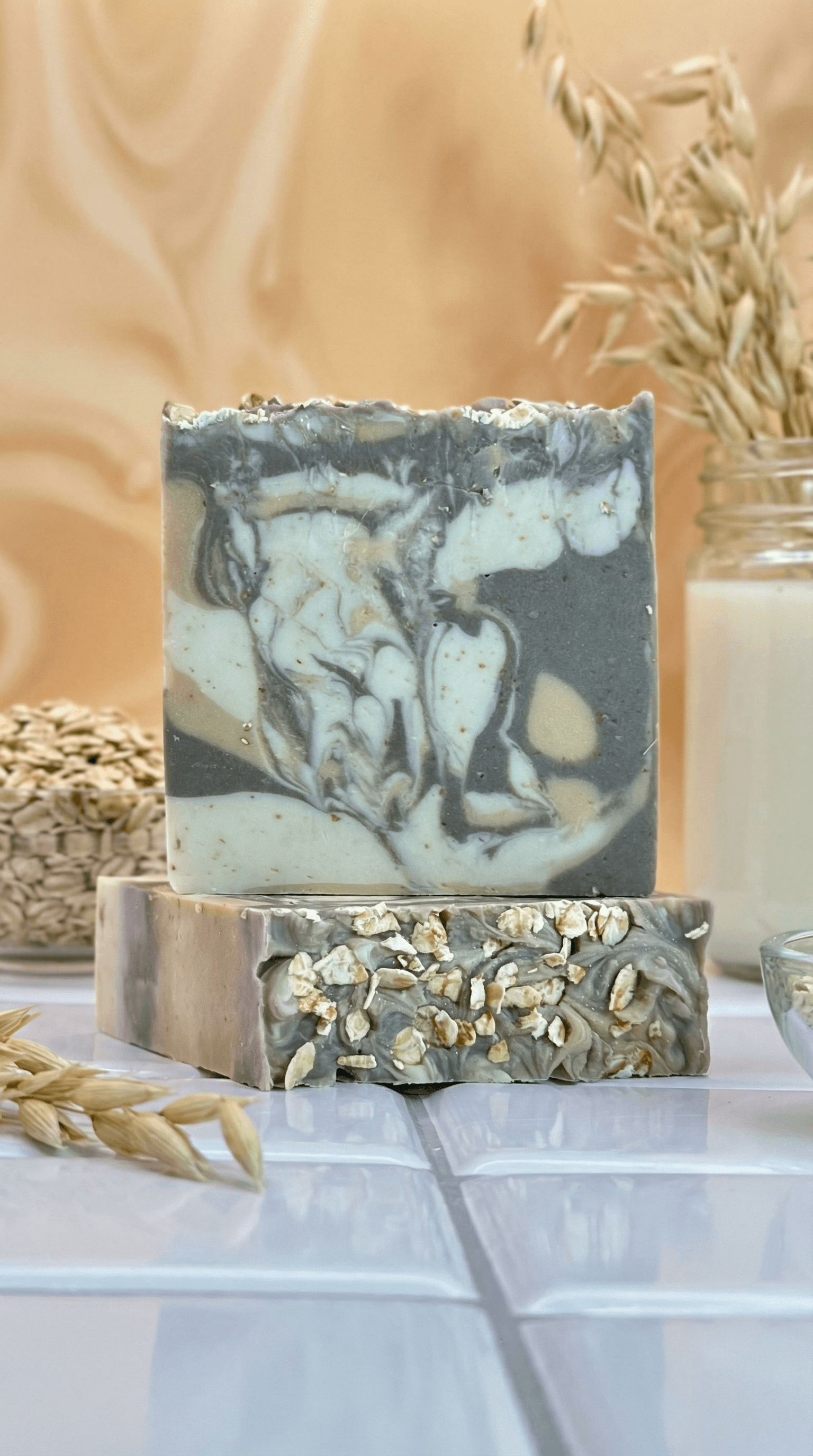 Two stacked handcrafted soap bars with marbled charcoal gray and cream swirls. The top bar has a textured surface, while the bottom bar is topped with whole oats against a natural backdrop.