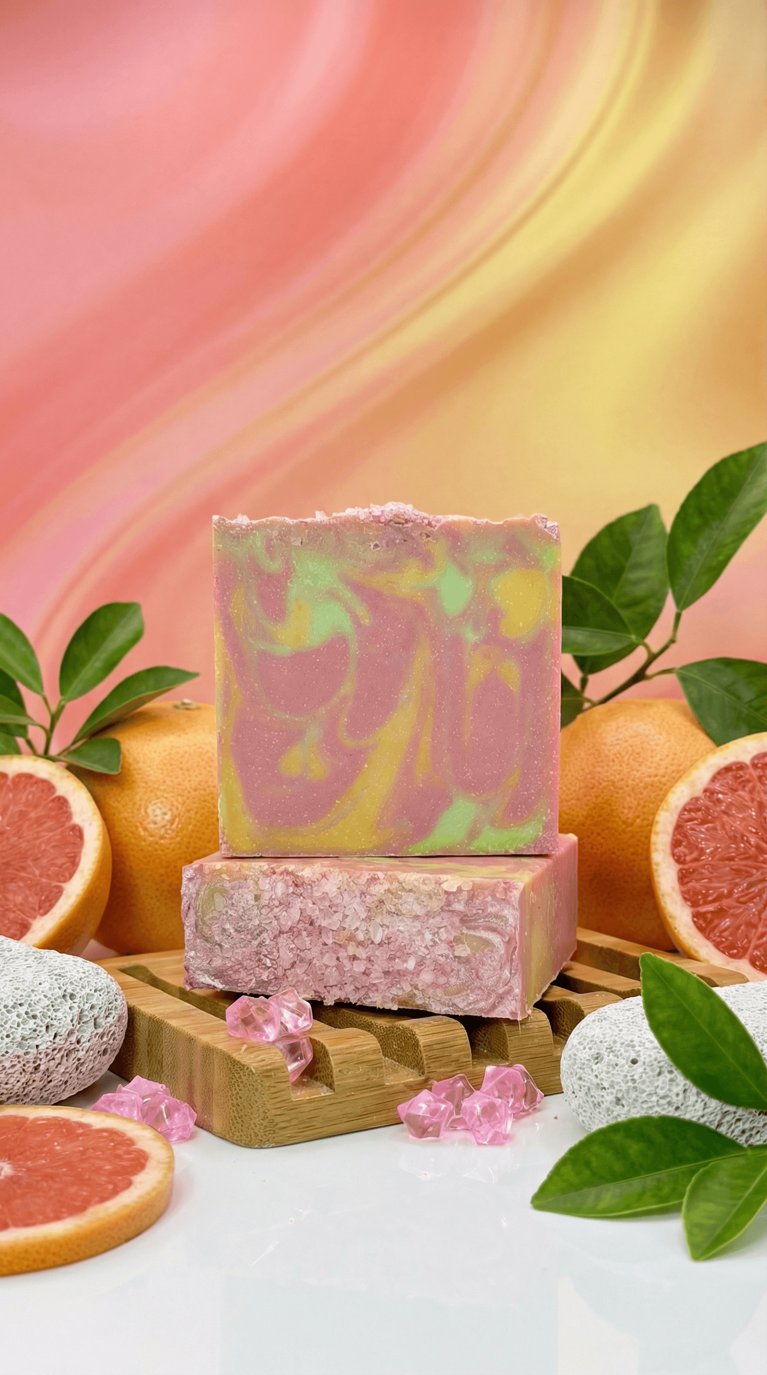 Two handcrafted soap bars stacked on bamboo, featuring pink and yellow-green swirls. Bottom bar shows visible pink salt crystals. Surrounded by fresh grapefruit slices and green leaves.