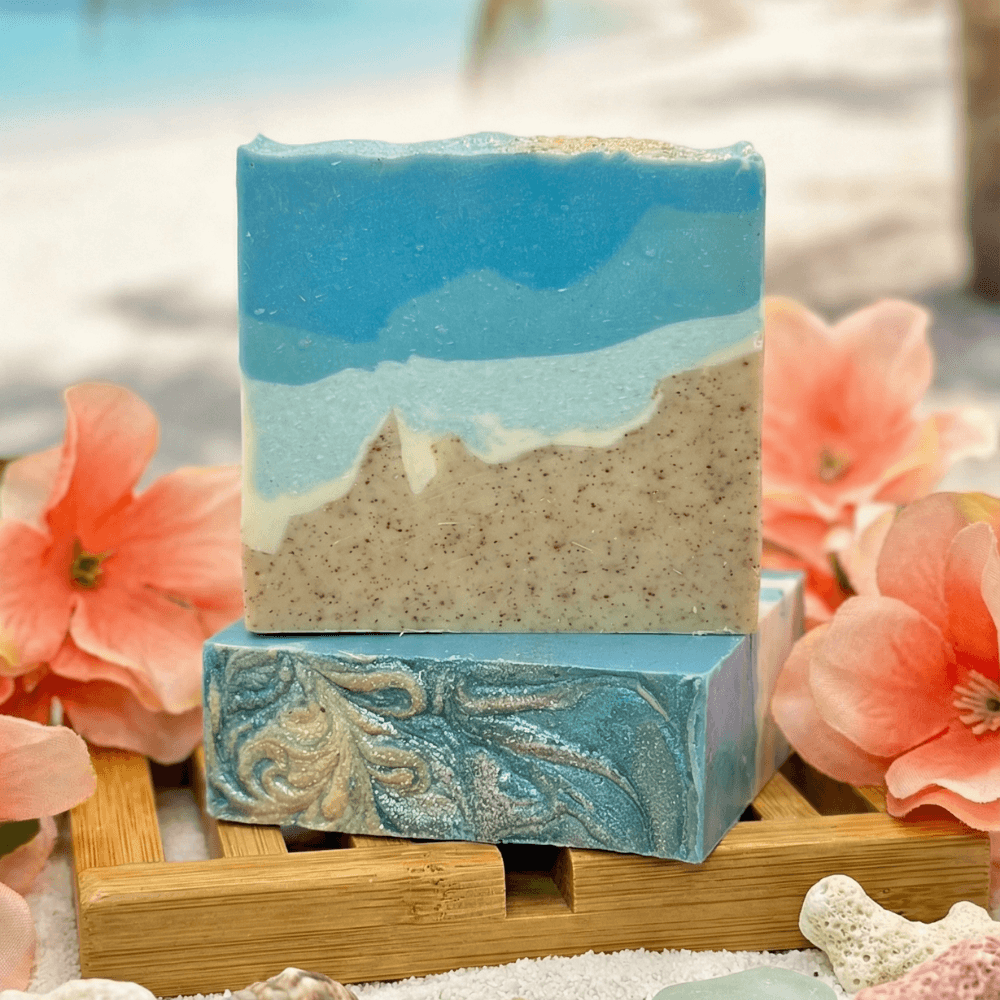 Two handcrafted soap bars stacked on bamboo. Top bar features layered blue ocean waves over tan speckled sand. Bottom bar shows swirled teal and gold design. Coral flowers frame the display.