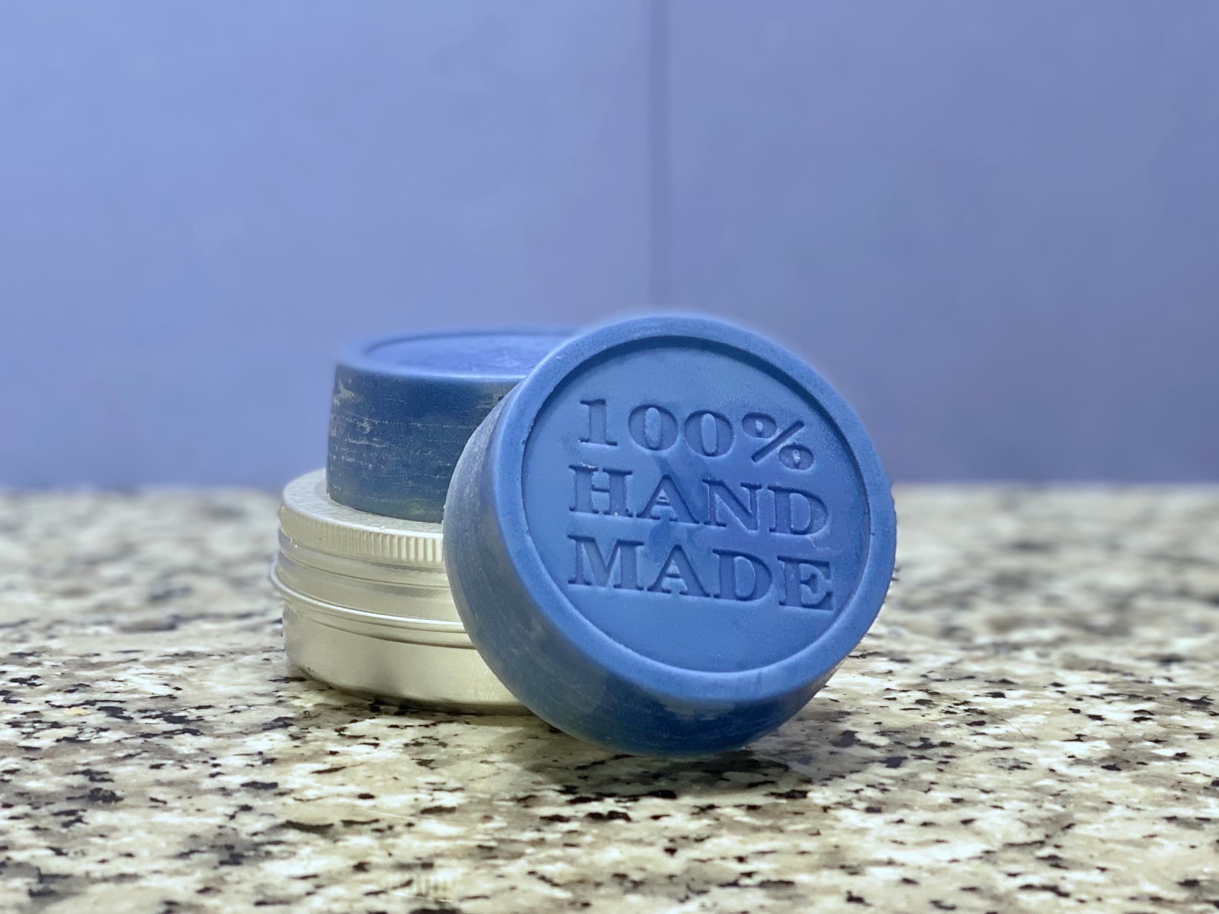 Blue circular solid lotion bar embossed with '100% HAND MADE' text, displayed on speckled counter with cream and dark colored containers behind it.