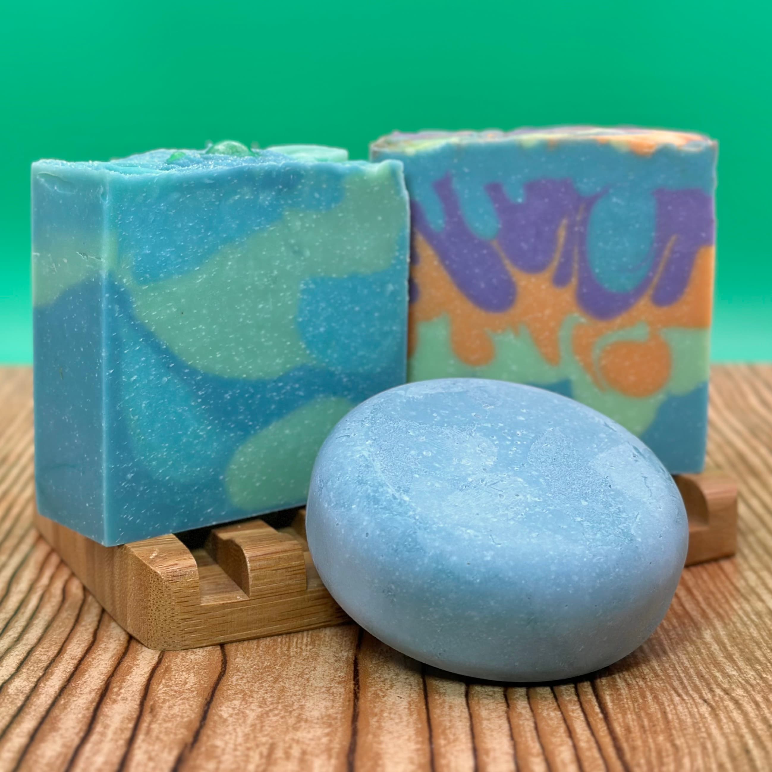 Three handcrafted soap bars in shades of blue and green on bamboo dishes. Two rectangular bars with swirled patterns and one smooth oval bar, displayed against a green background.