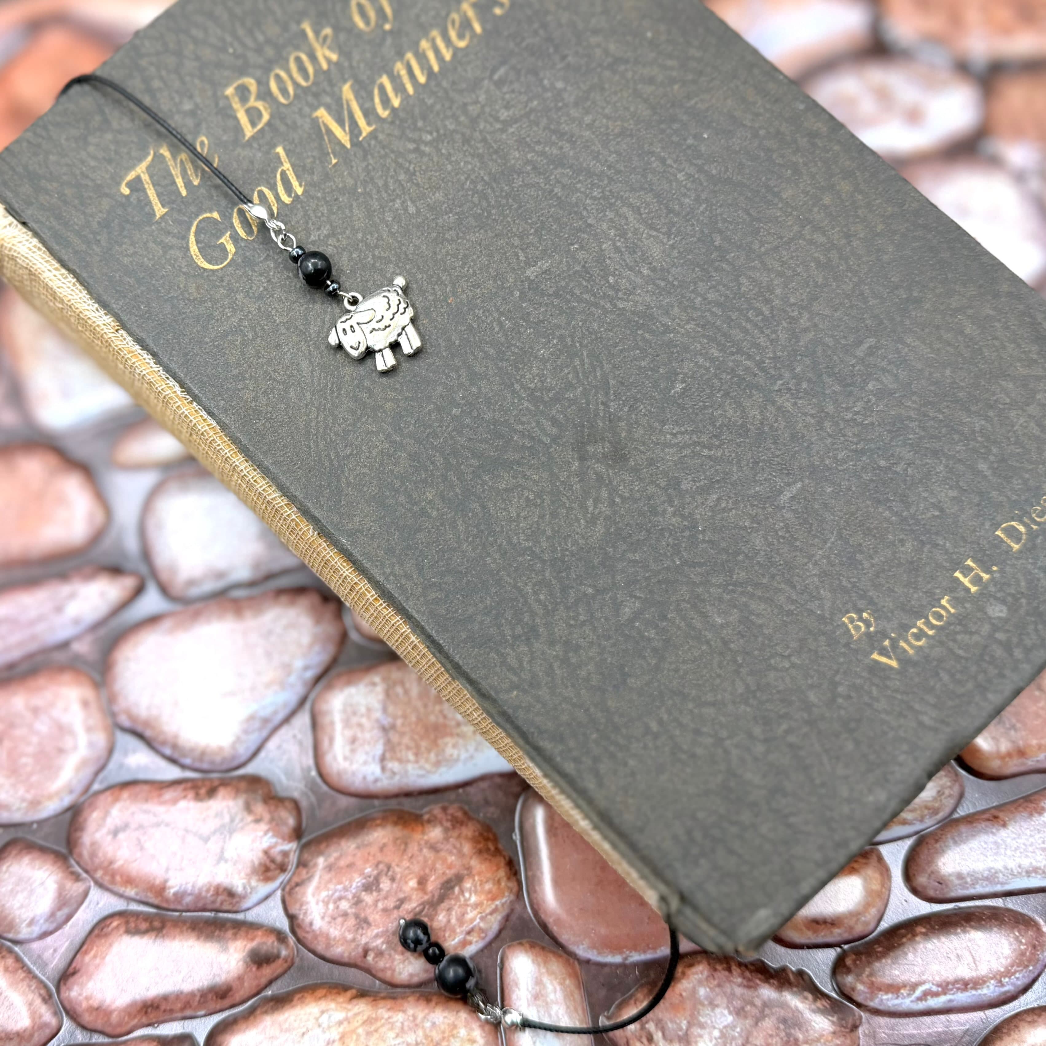 Gray hardcover book titled 'The Book of Good Manners' with gold lettering, featuring a decorative elephant charm bookmark with black beads resting on its cover against a river stone background.