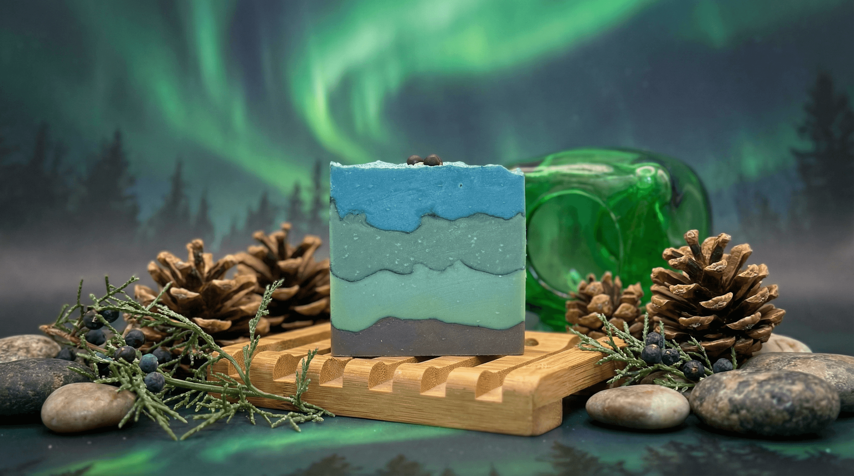 Layered bar soap in ombre shades of charcoal gray, seafoam green, mint, and sky blue with wavy horizontal layers. Displayed on bamboo tray with pine cones, juniper sprigs, river stones, and green glass bottle against northern lights backdrop.