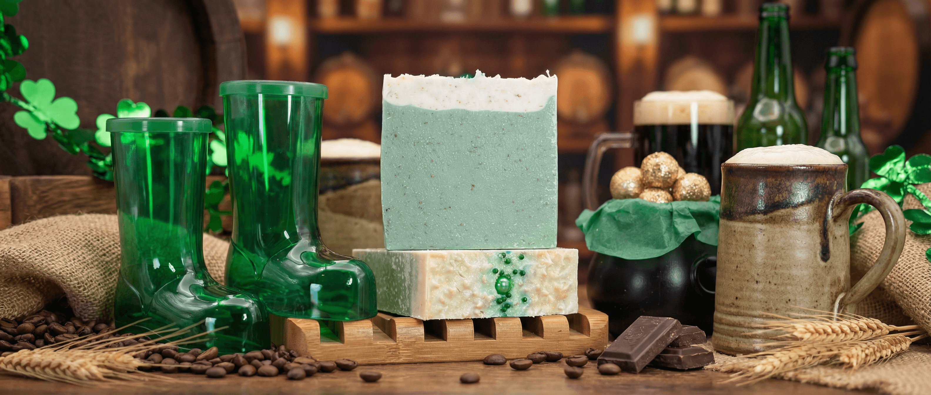 Two handcrafted soap bars on a wooden dish: one large mint-green bar with white foam topping, one smaller cream-colored bar with green shamrock embed. Surrounded by green beer glasses, coffee beans, and Irish pub decor.