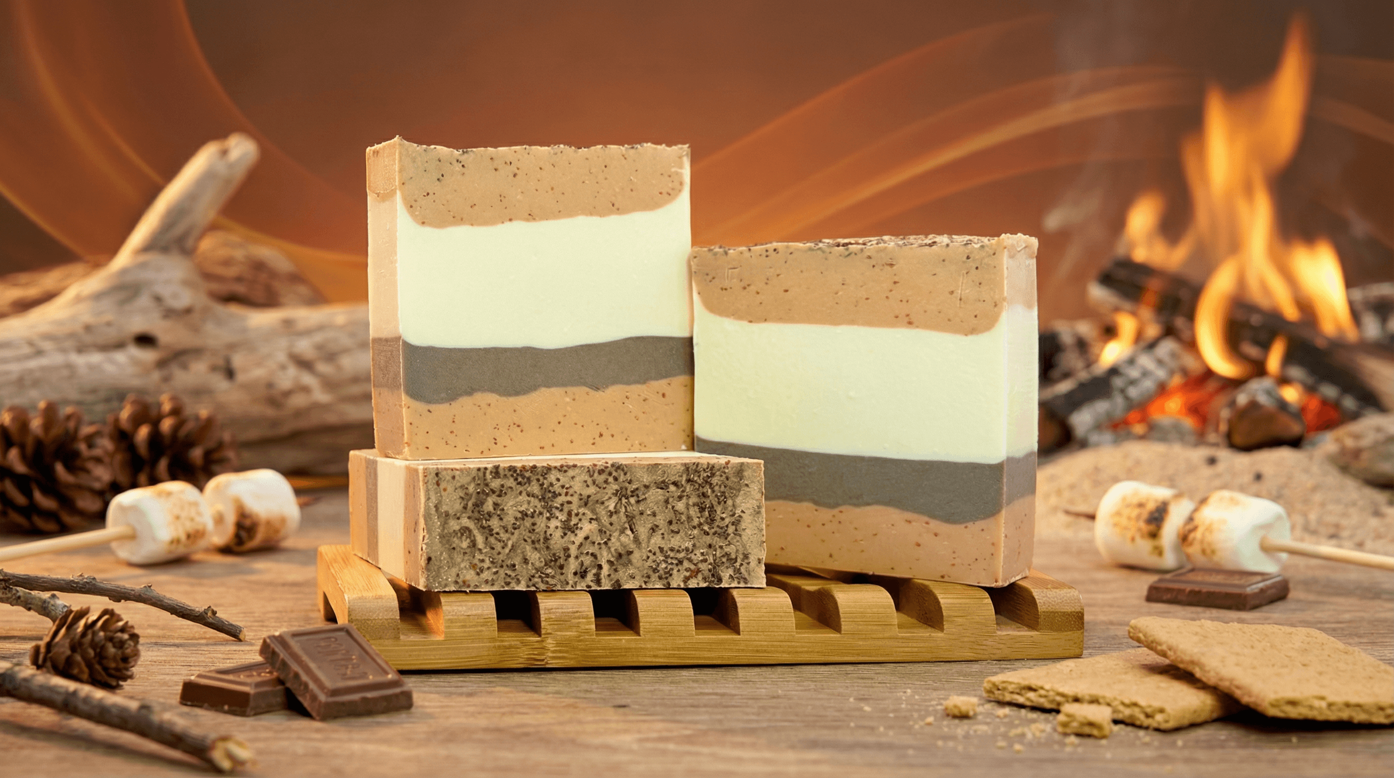 Three bars of layered handcrafted soap in tan, cream, and sage green stripes with speckled texture, displayed on wooden soap dish. Styled with s'mores ingredients, pine cones, and fireplace.