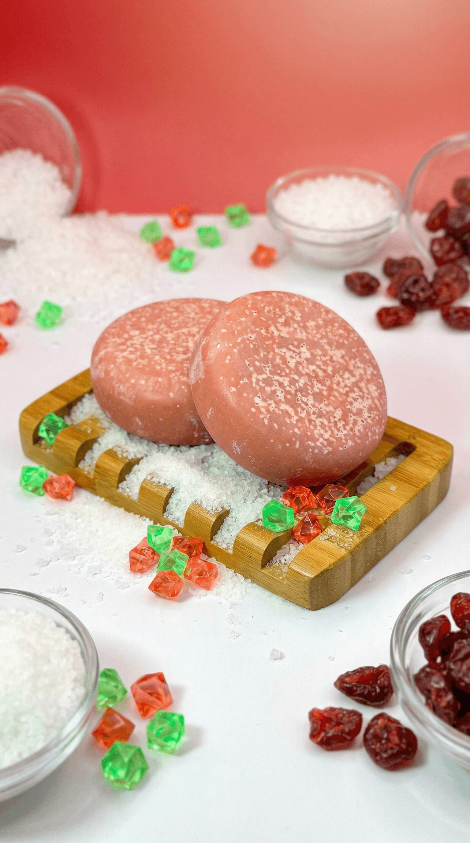 Two peachy-pink oval soap bars with coarse white salt crystals on surface, displayed on wooden soap dish surrounded by scattered white salt, dried cranberries, and colorful candy-like gems.