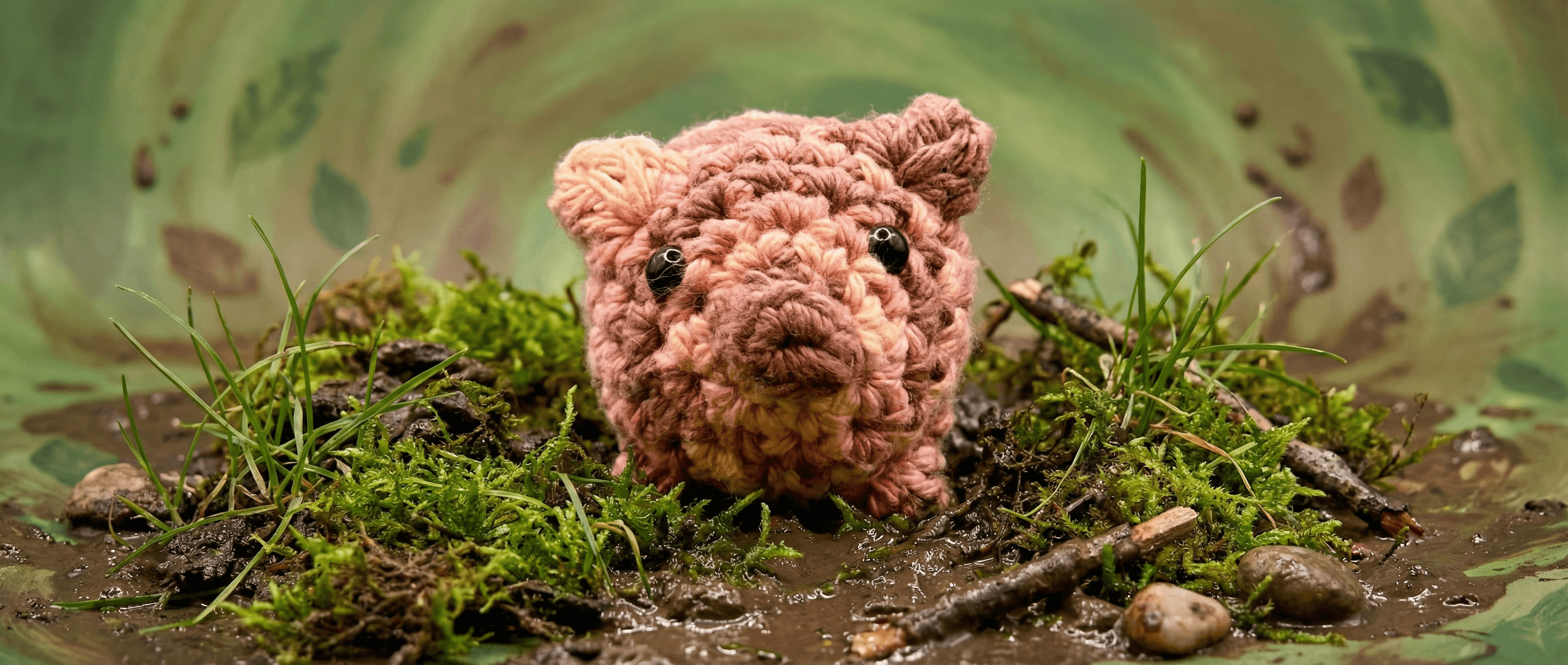 Pink and brown variegated crochet pig sachet with small black bead eyes, sitting on moss-covered wood and twigs against a soft green floral background.