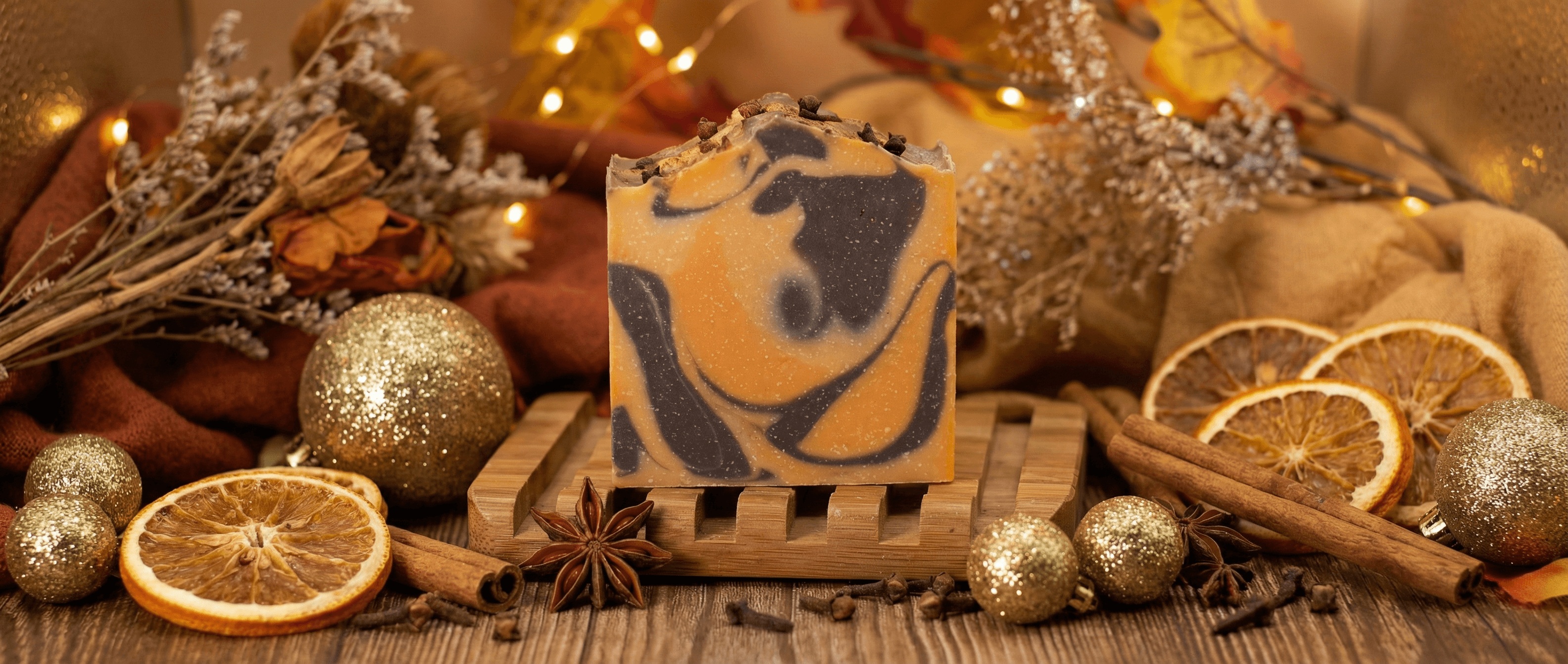 Handcrafted soap bar with orange and dark brown swirled marbling, topped with whole cloves, displayed on wooden soap dish surrounded by dried orange slices, cinnamon sticks, star anise, and gold ornaments.