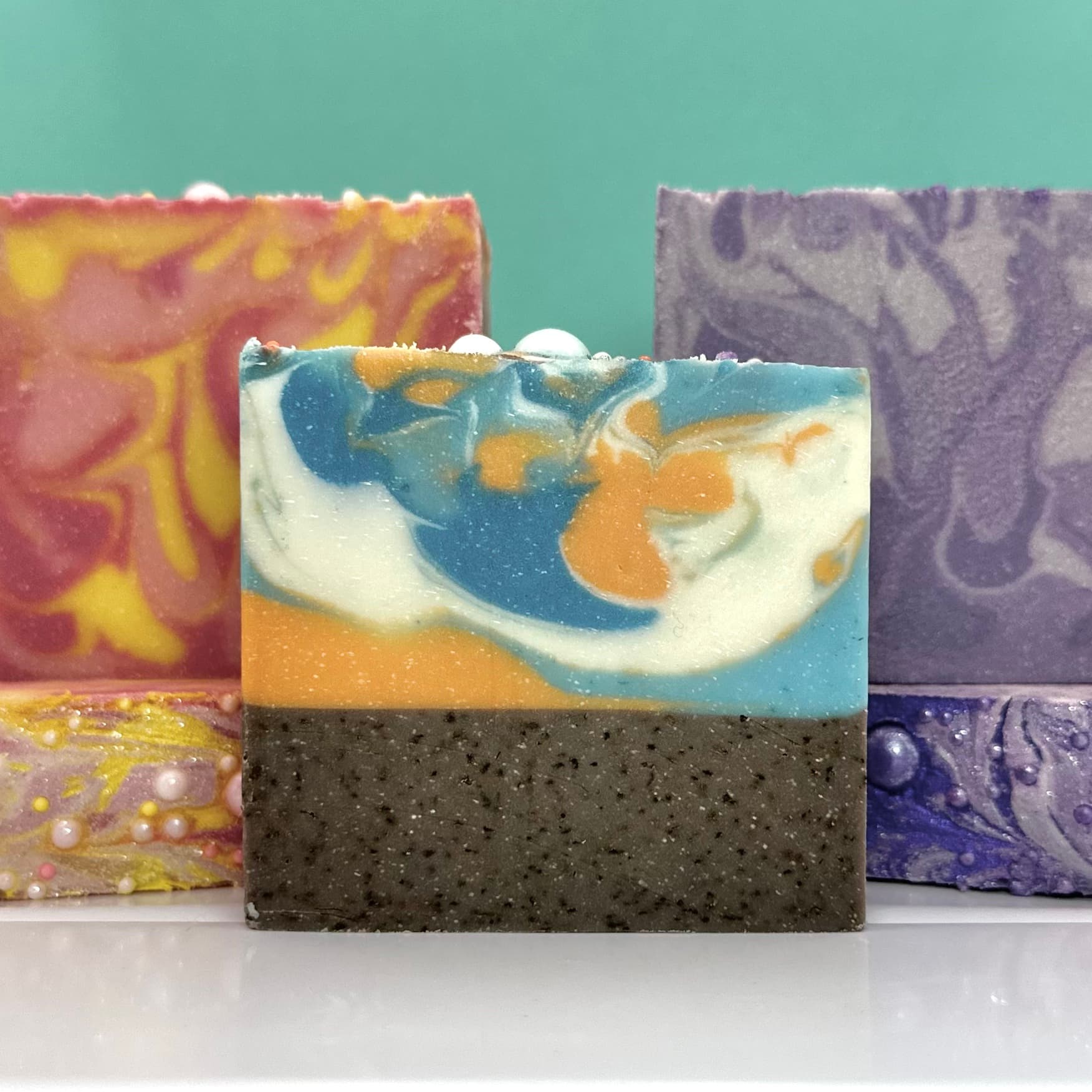 Five handcrafted soap bars with swirled patterns in various color combinations: pink-yellow, blue-orange-white with dark speckled base, and purple. Each bar displays marbled designs on their surfaces.