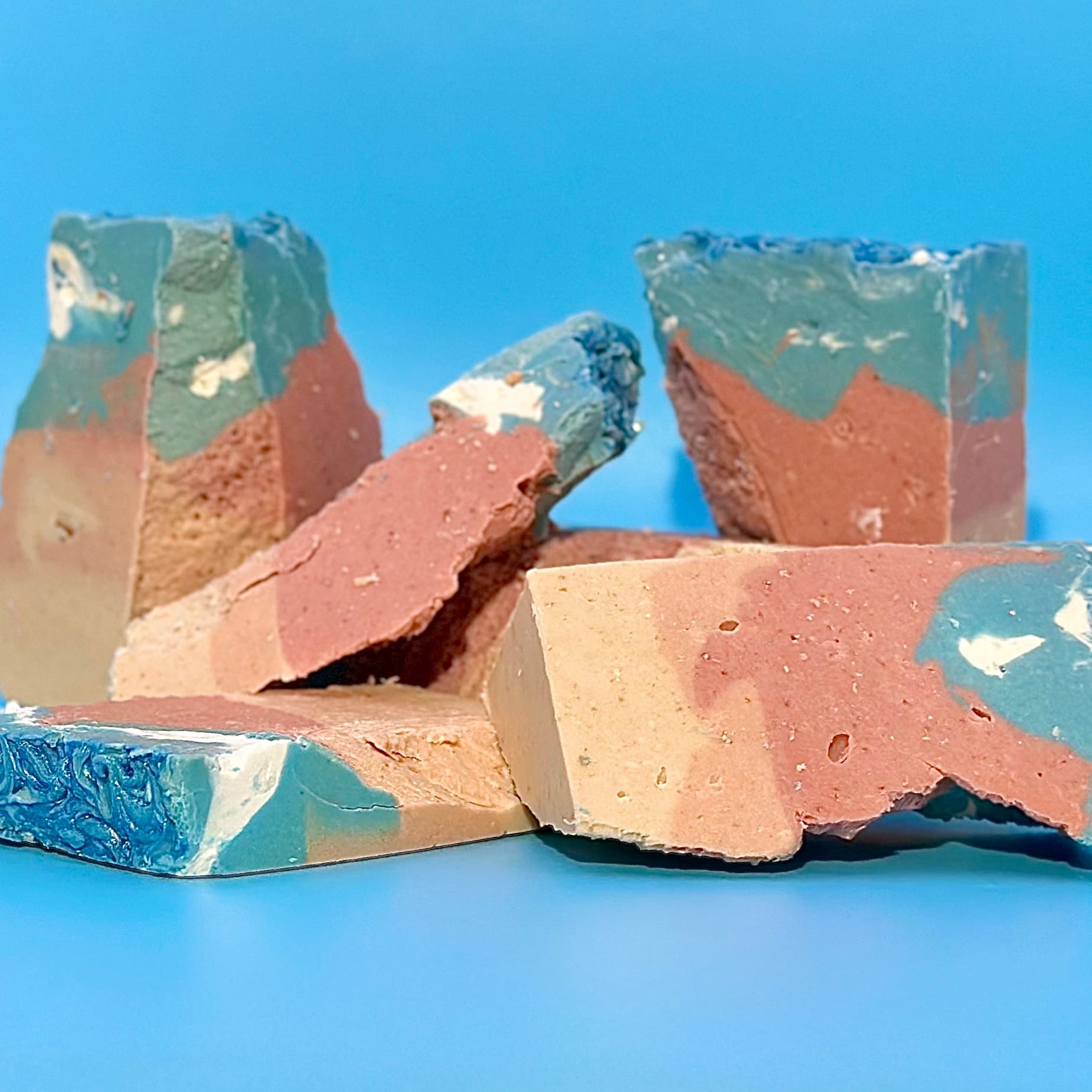 Several broken soap pieces displaying swirled layers of teal blue, coral pink, cream, and tan colors with visible exfoliating specks throughout, arranged on a bright blue background.