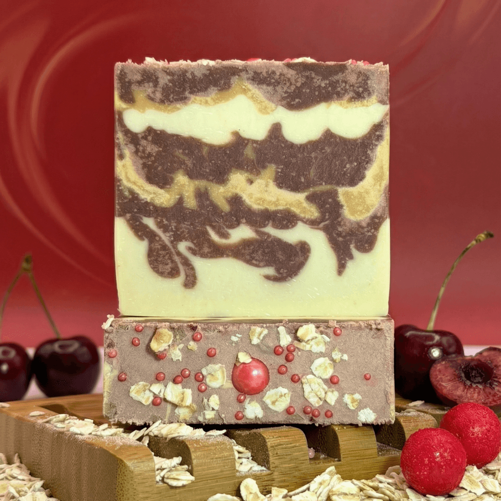 Two stacked handmade soap bars on a wooden soap dish. Top bar features cream and brown swirled layers. Bottom bar is brown with oat pieces, red spherical embeds, and a central candy.