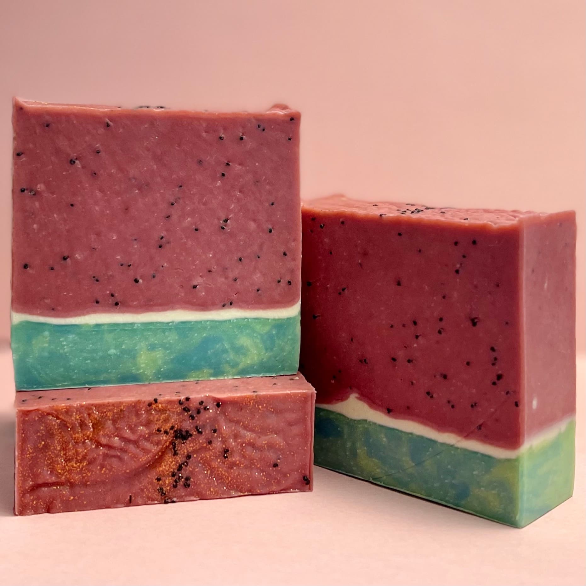 Three rectangular bars with pink watermelon-colored tops speckled with black poppy seeds, white middles, and teal-green rinds layered to resemble watermelon slices, arranged on a pink surface.