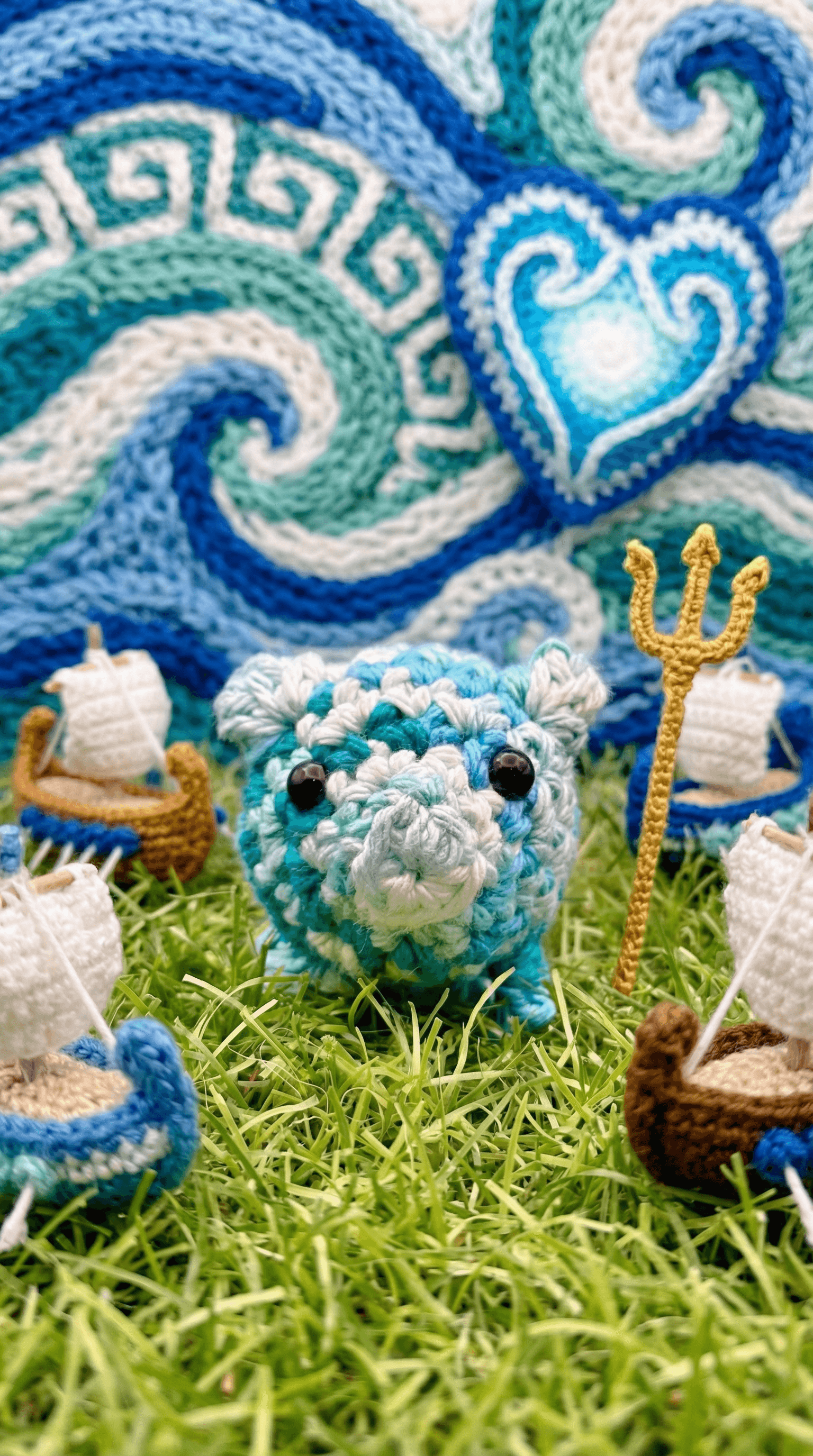 Turquoise and white striped crocheted pig wearing a golden trident, positioned on artificial grass with ocean wave tapestry backdrop and small white sheep scattered around.