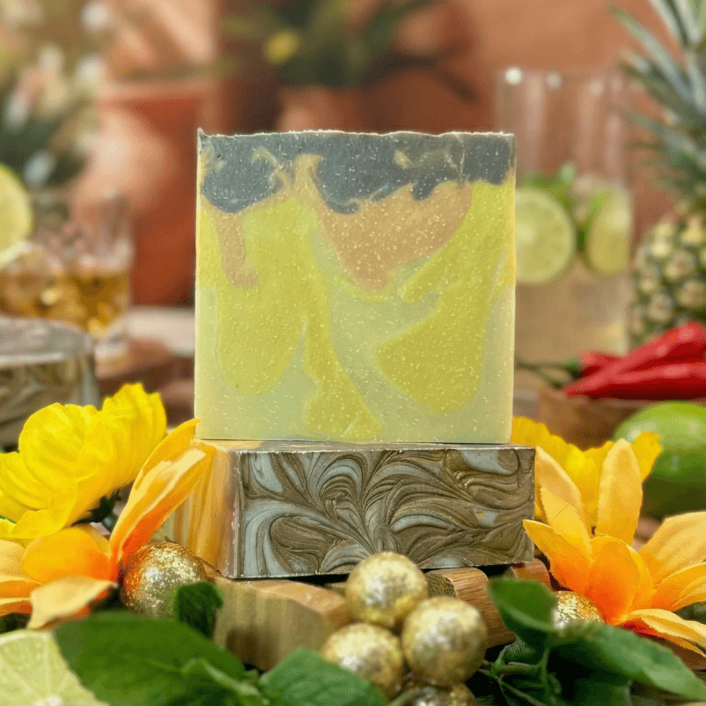 Two stacked handmade soap bars with marbled swirls in yellow, grey, and olive green tones, displayed on a wooden holder surrounded by bright yellow flowers and golden ornaments.