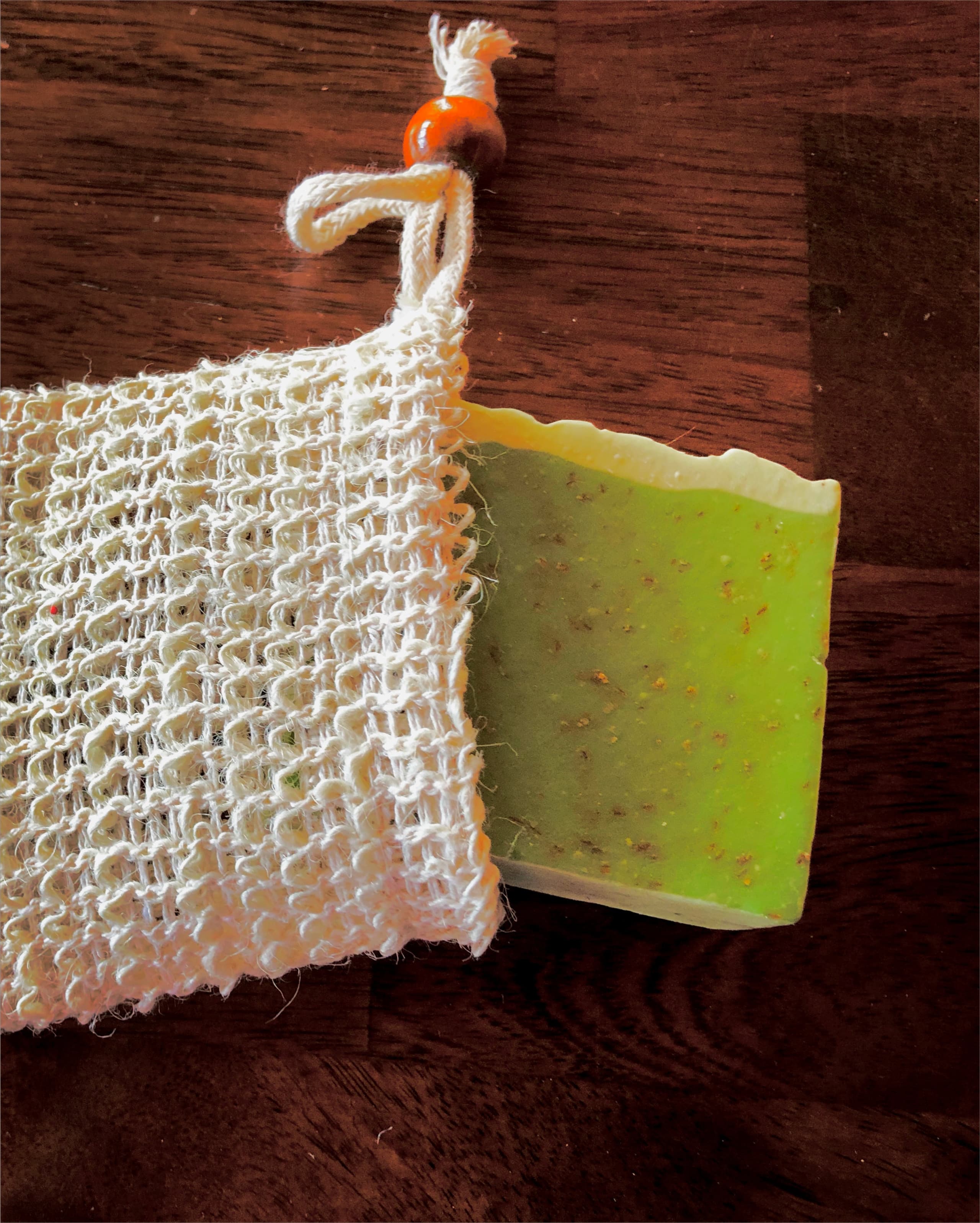 Natural sisal mesh pouch with drawstring beside a lime green rectangular soap bar speckled with orange and green botanical pieces on a wooden surface.