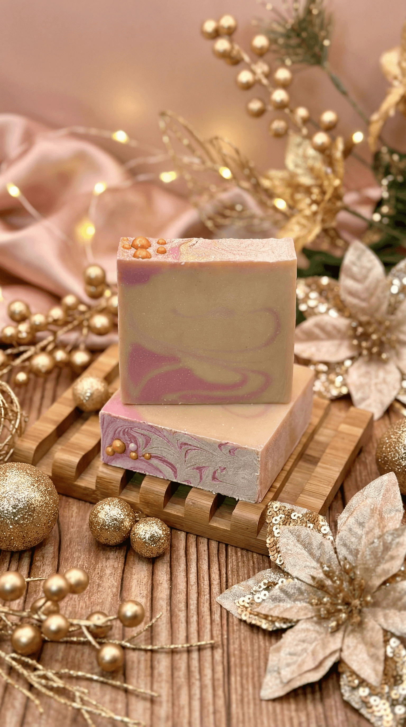 Two bars of handcrafted soap with creamy beige and dusty purple swirls, topped with gold biodegradable glitter beads, displayed on a bamboo soap dish surrounded by golden holiday decorations.