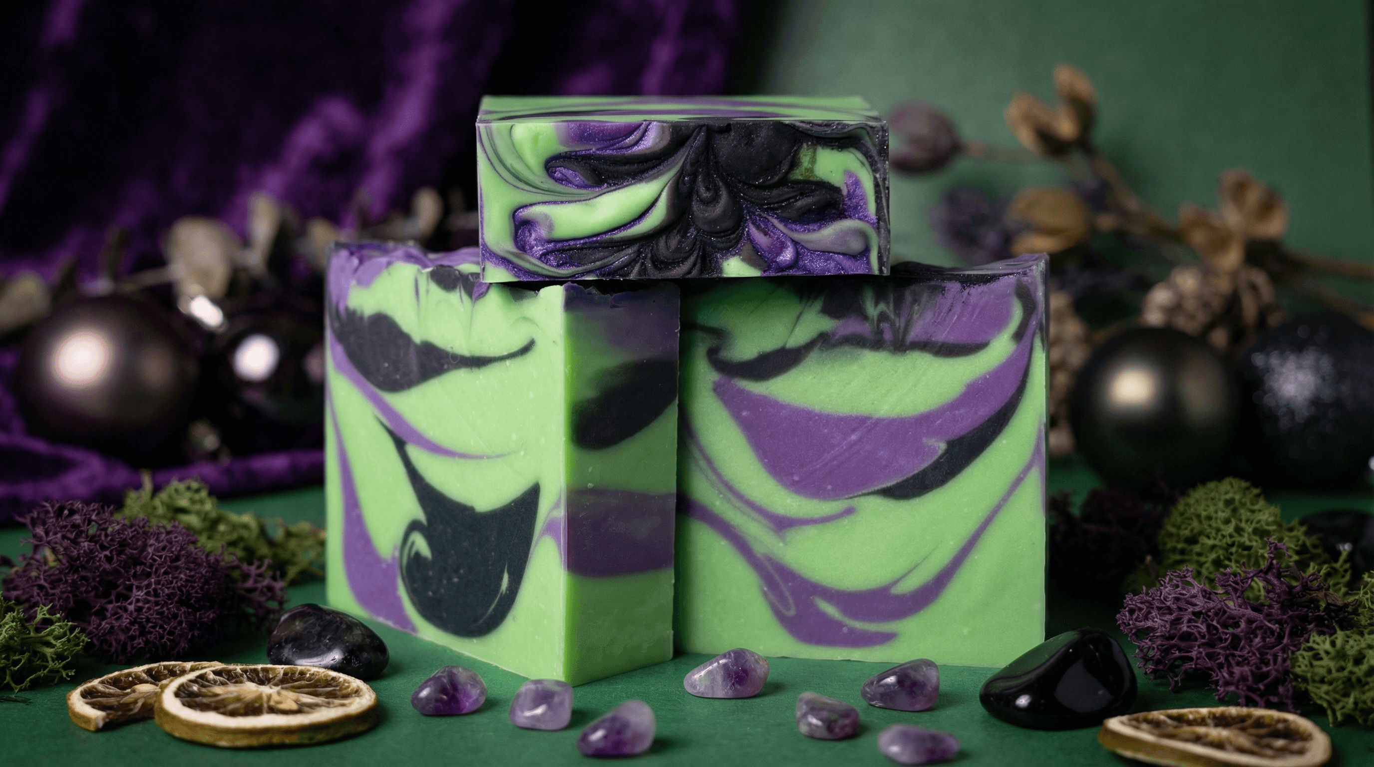 Three bars of handcrafted soap with bold lime green base and purple and black marbled swirls, displayed on moss with dried citrus slices and purple amethyst stones.