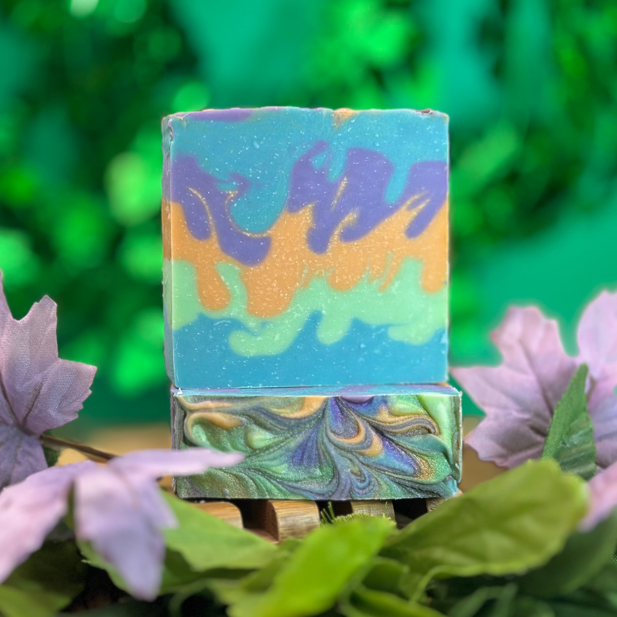 Two stacked handmade soap bars with swirled marbling in turquoise, purple, gold, and green tones, displayed on a wooden soap dish surrounded by purple flowers and greenery.