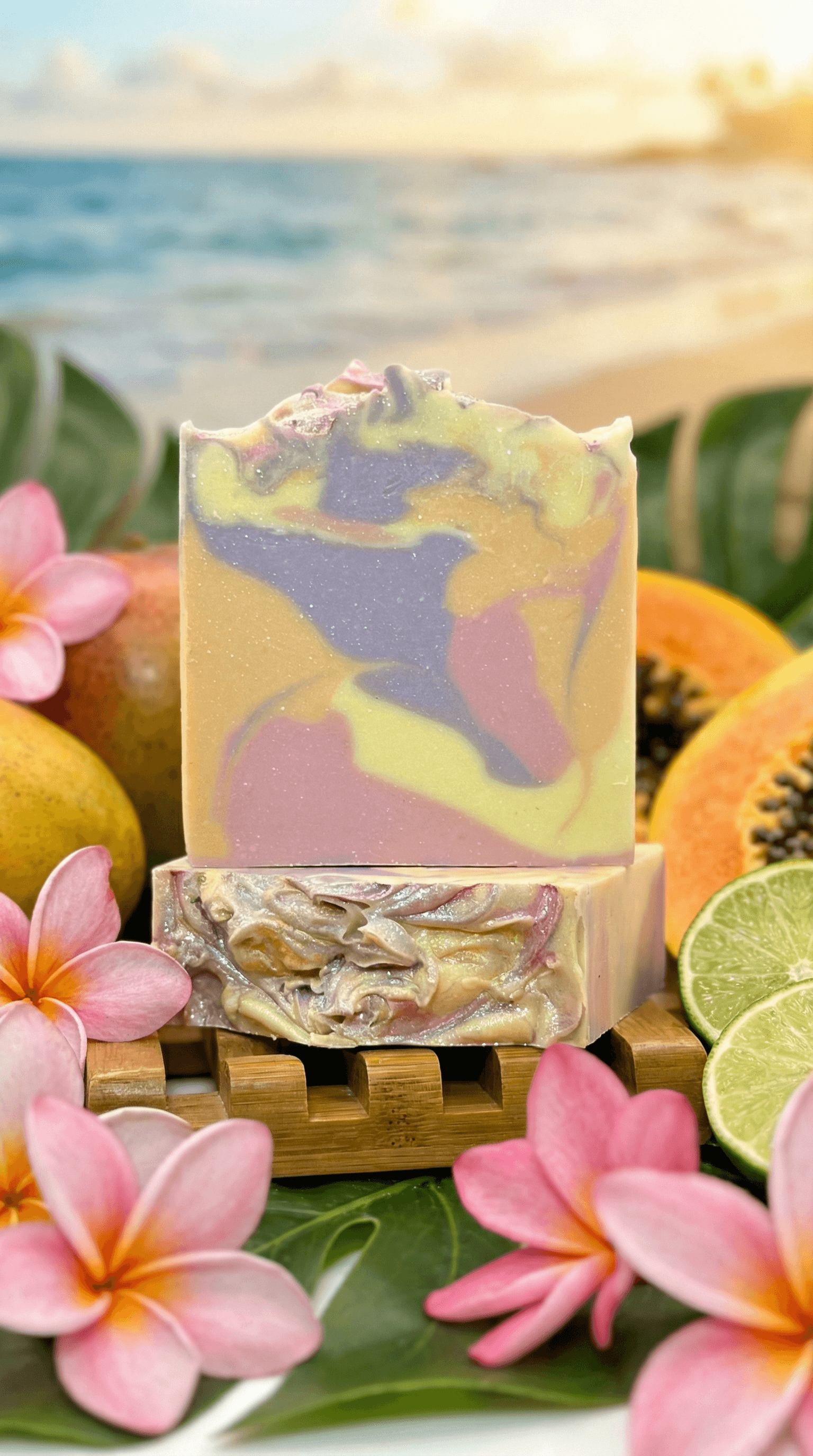 Two bars of handcrafted soap with pastel swirls of yellow, lavender, pink, and green, displayed on a bamboo tray surrounded by tropical plumeria flowers, papaya, mango, and lime against a beach backdrop.