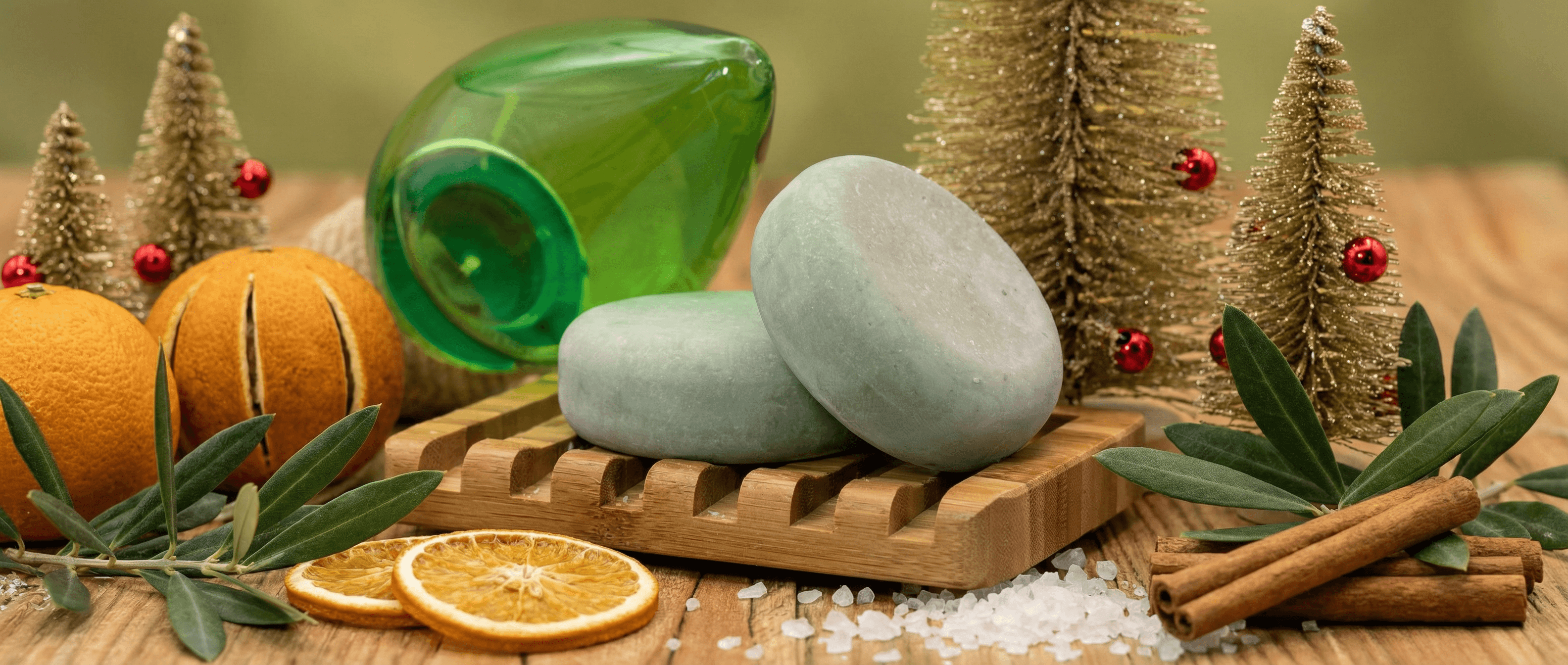 Two pale gray-blue oval soap bars with smooth matte finish rest on a wooden soap dish, surrounded by fresh oranges, cinnamon sticks, sea salt, olive branches, and miniature bottle brush trees with red ornaments in a rustic holiday scene.