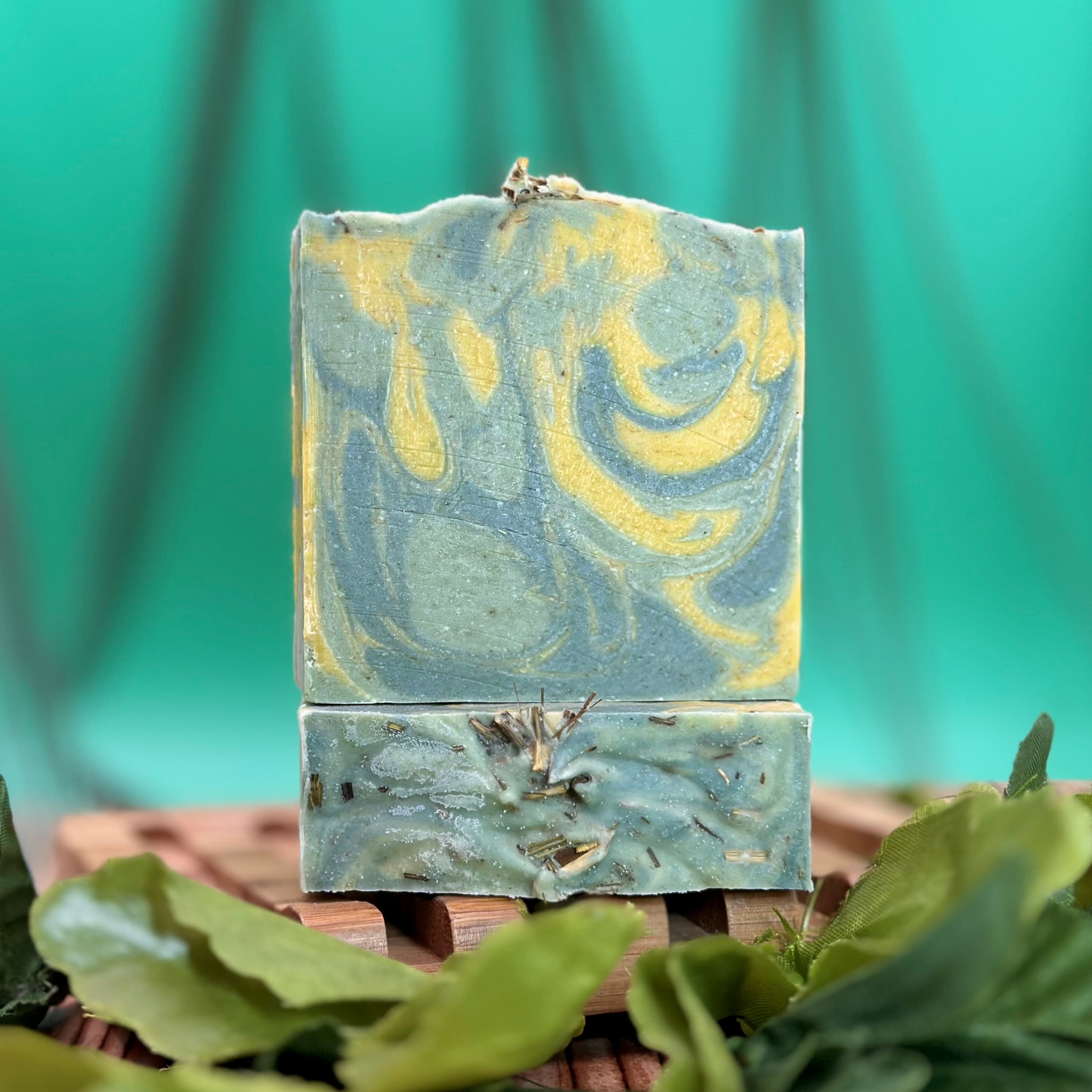 Two stacked handmade soap bars in dusty blue-green with yellow swirls on top bar, visible herb pieces in bottom bar. Displayed on wooden slat surface with green leaves.