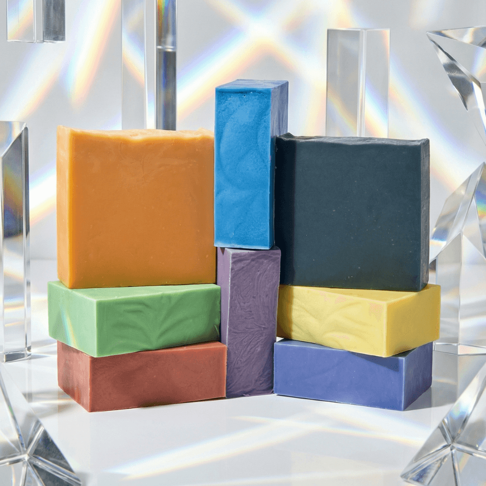 Eight rectangular soap bars in rainbow colors plus black, stacked in a geometric arrangement against a prismatic background with clear acrylic elements and rainbow light refractions.