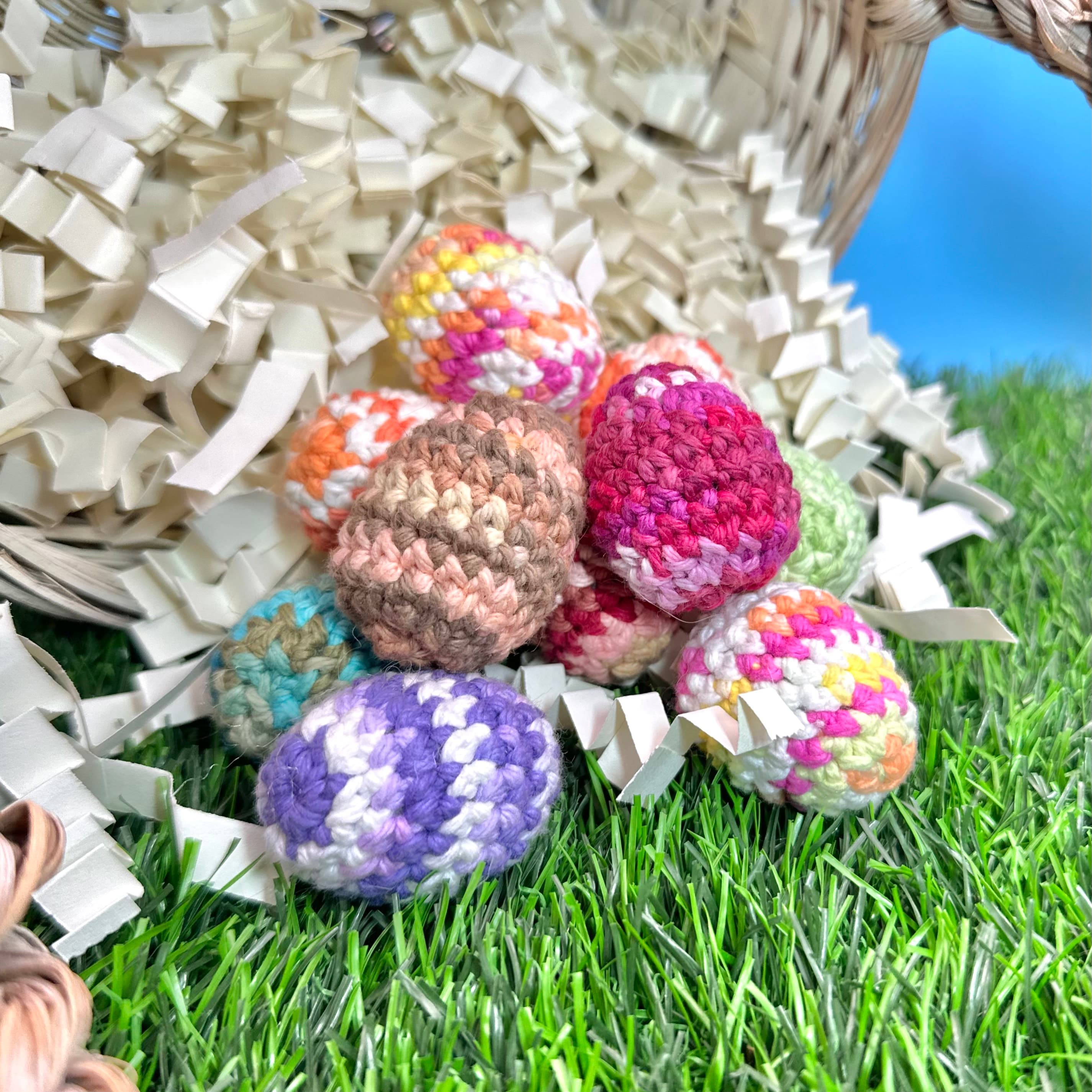 Seven colorful crocheted eggs in various patterns spilling from a wicker basket onto artificial grass with white paper shred. Eggs feature pink, purple, brown, and multicolor yarn.
