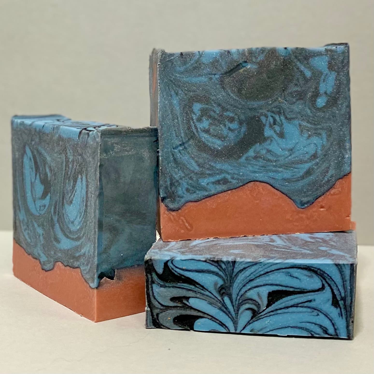 Three rectangular soap bars with marbled blue and charcoal swirls on top and terracotta-colored base. The bars show an organic, handcrafted swirl pattern throughout.