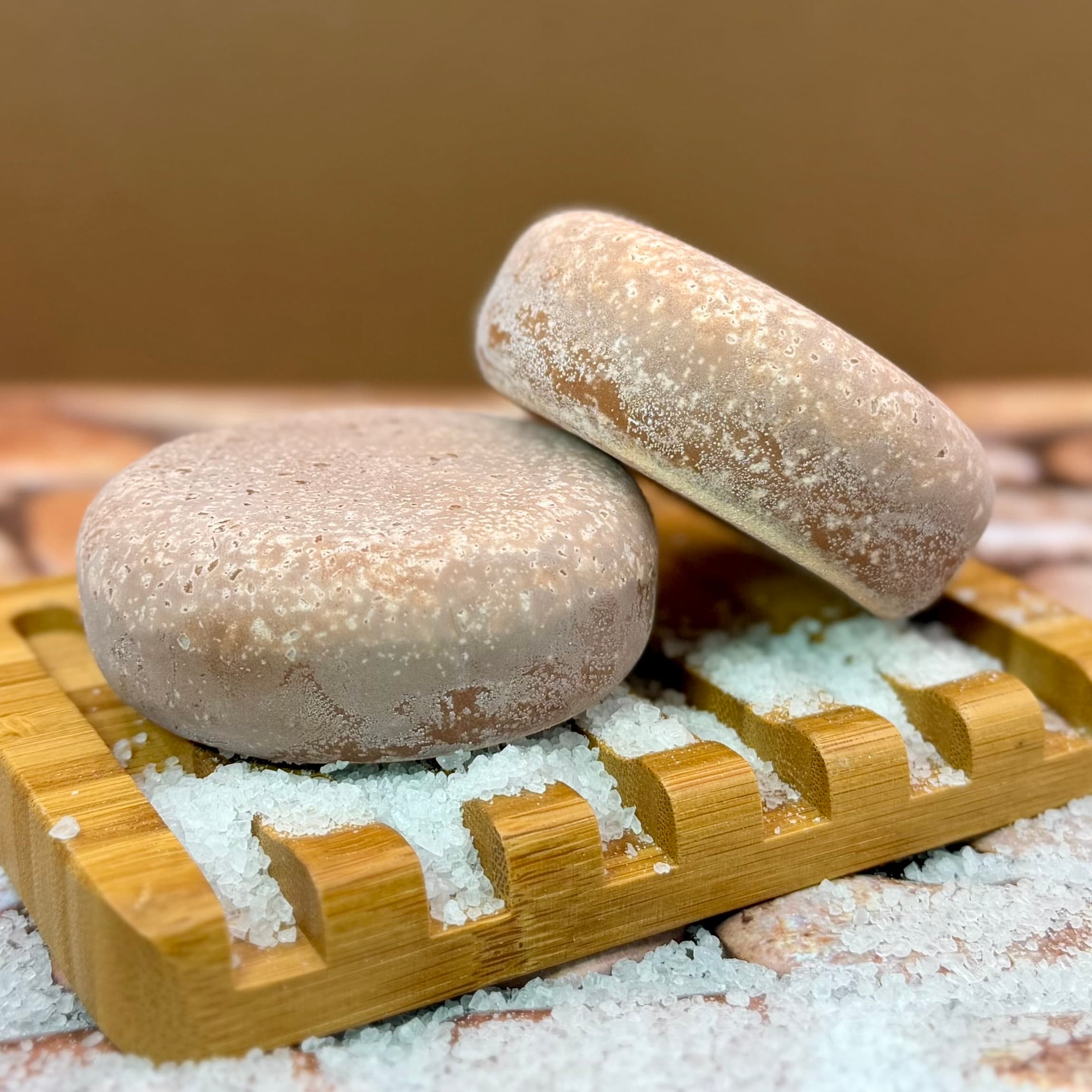 Two round, frosted beige soap bars stacked on a bamboo dish with coarse white sea salt crystals scattered around. The bars have a textured, salt-crusted surface.