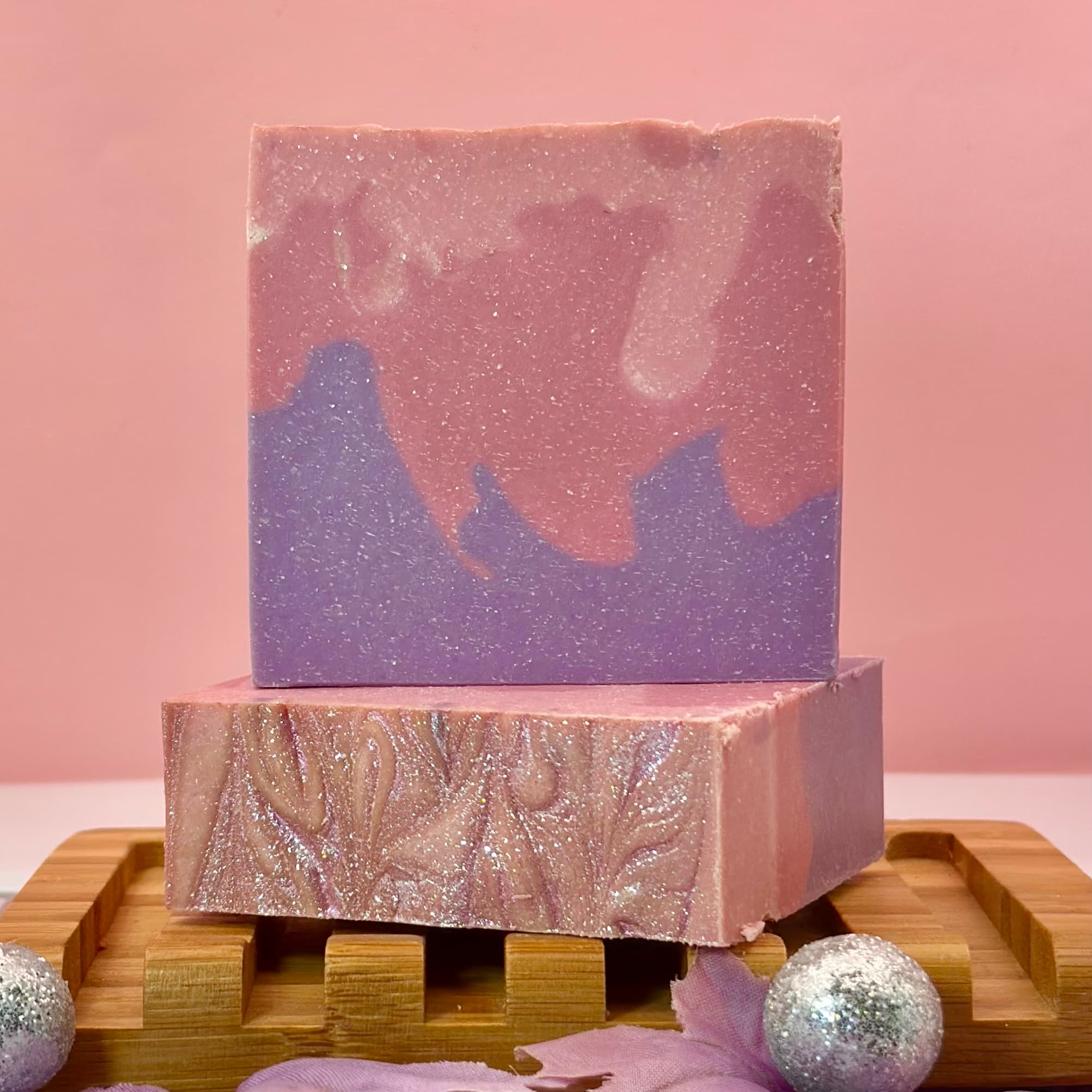 Two handcrafted soap bars on a wooden soap dish with pink and purple swirled colors, topped with a sparkly glittered surface. The front bar features an embossed floral design.