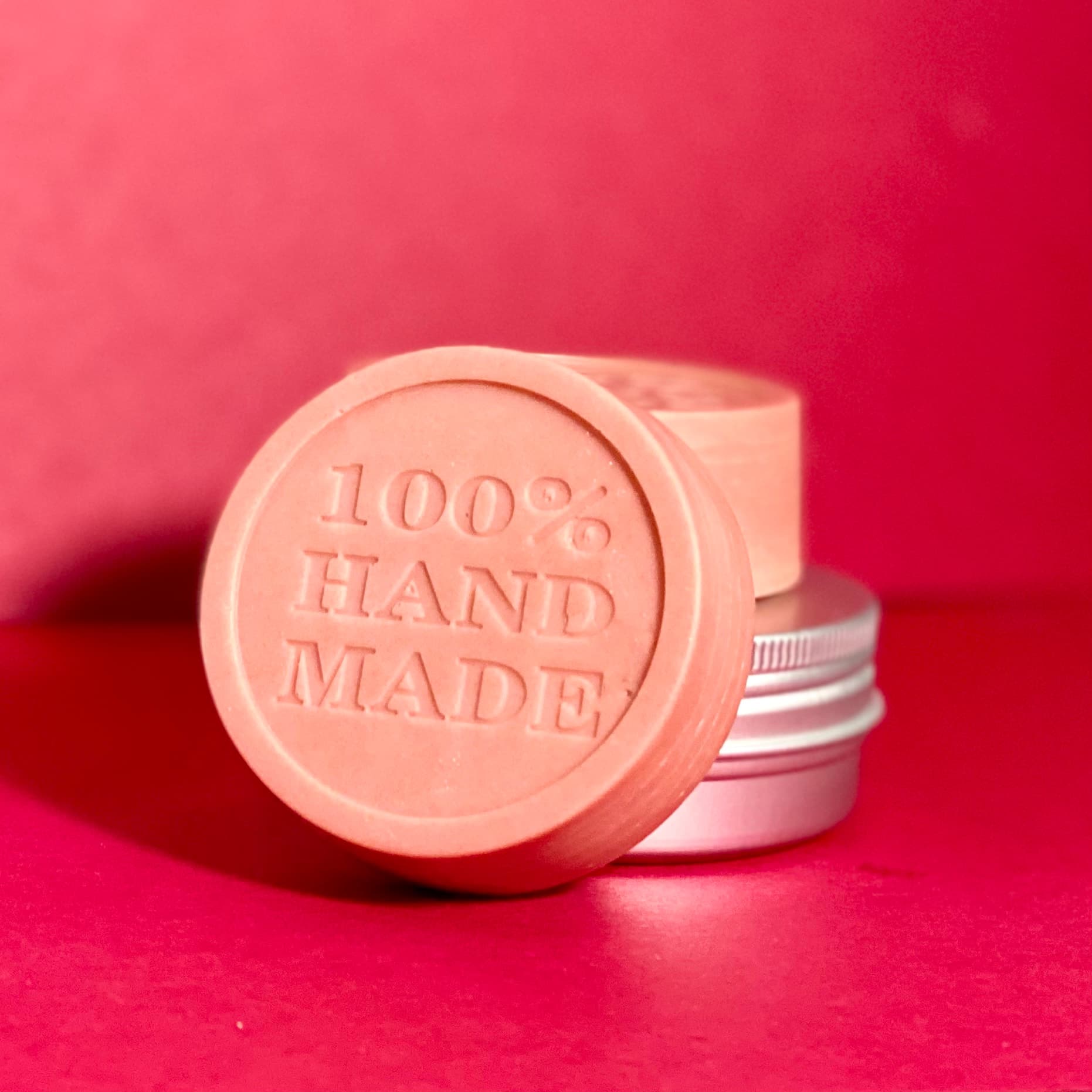 Peachy pink round solid lotion bar embossed with '100% HAND MADE' text, displayed with silver and white tins against vibrant pink background.