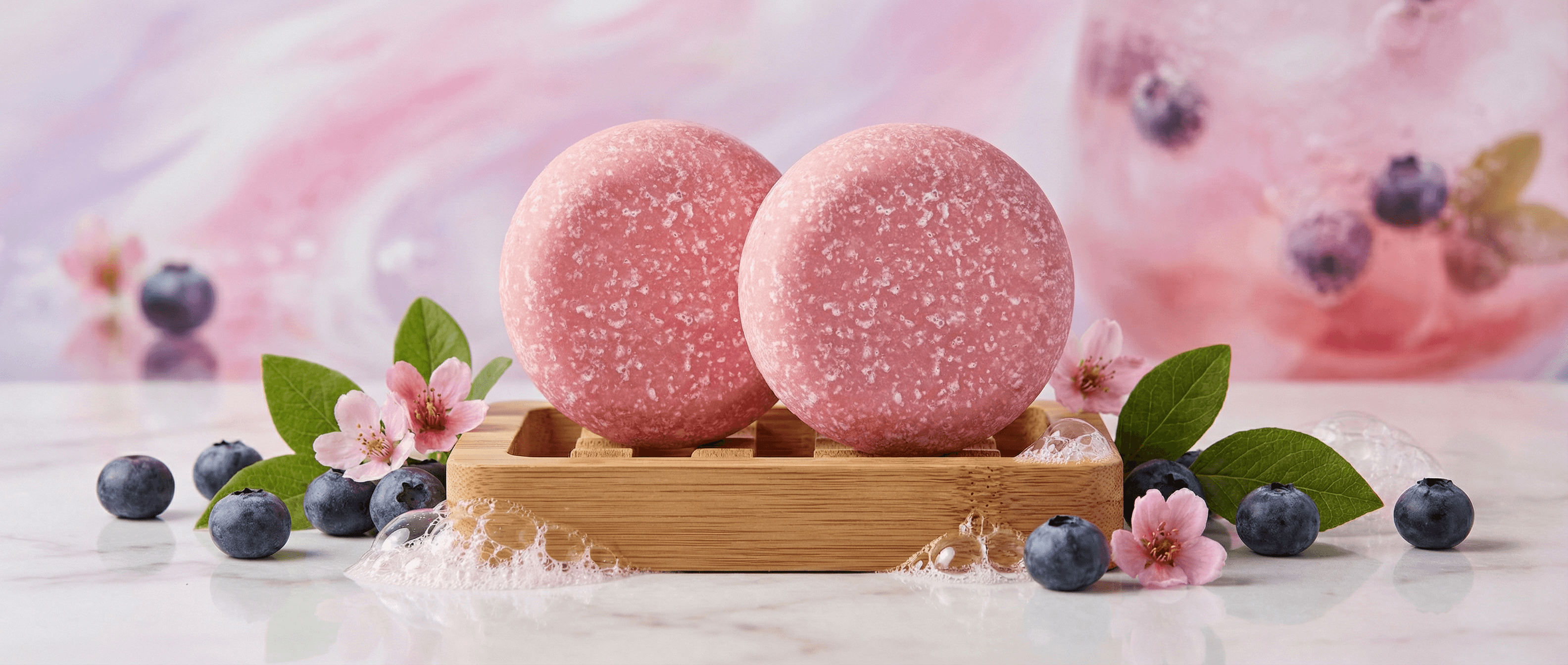 Two round pink sea salt scrub bars with crystalline texture on a bamboo dish, surrounded by fresh blueberries, pink blossoms, and green leaves on a white surface.
