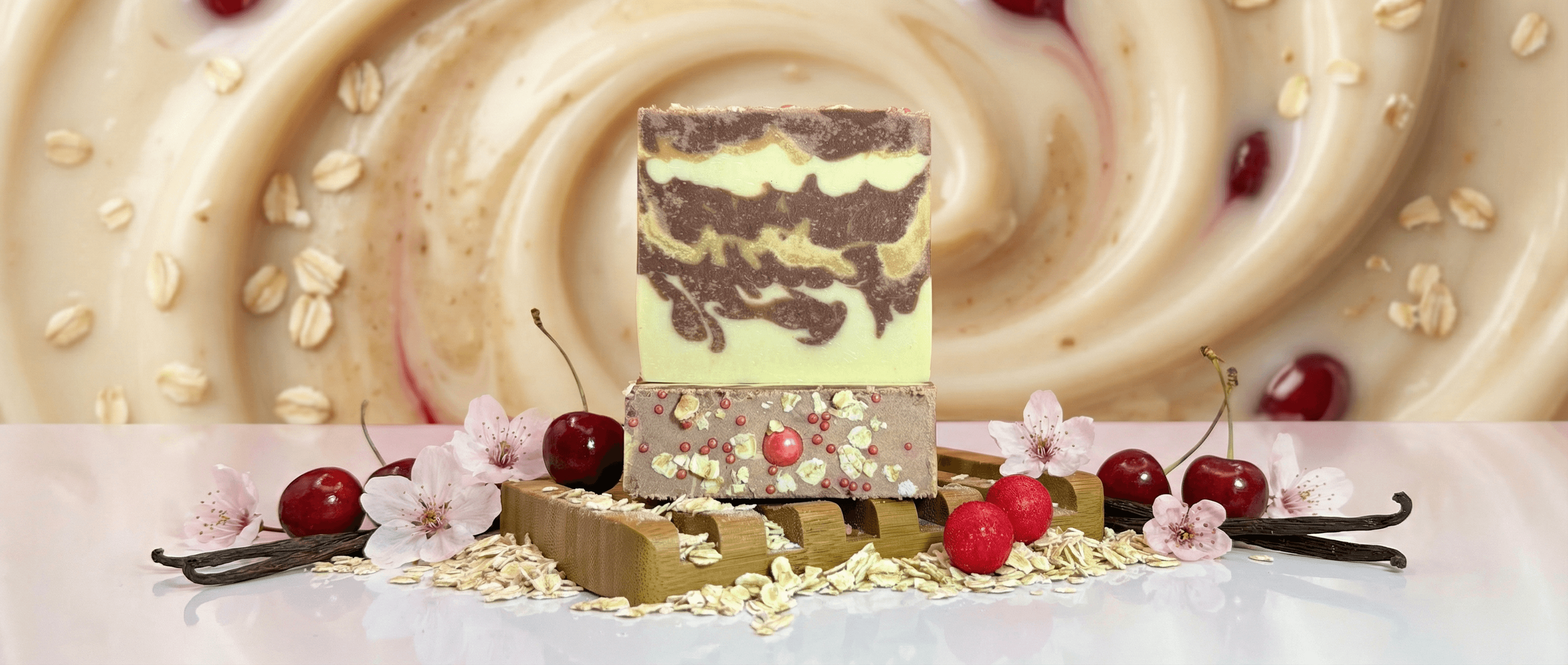 Two bars of handcrafted soap stacked on a wooden soap dish. Top bar is pale green with dark brown swirls. Bottom bar is speckled with oats and pink cranberries, surrounded by cherries, vanilla beans, and cherry blossoms.