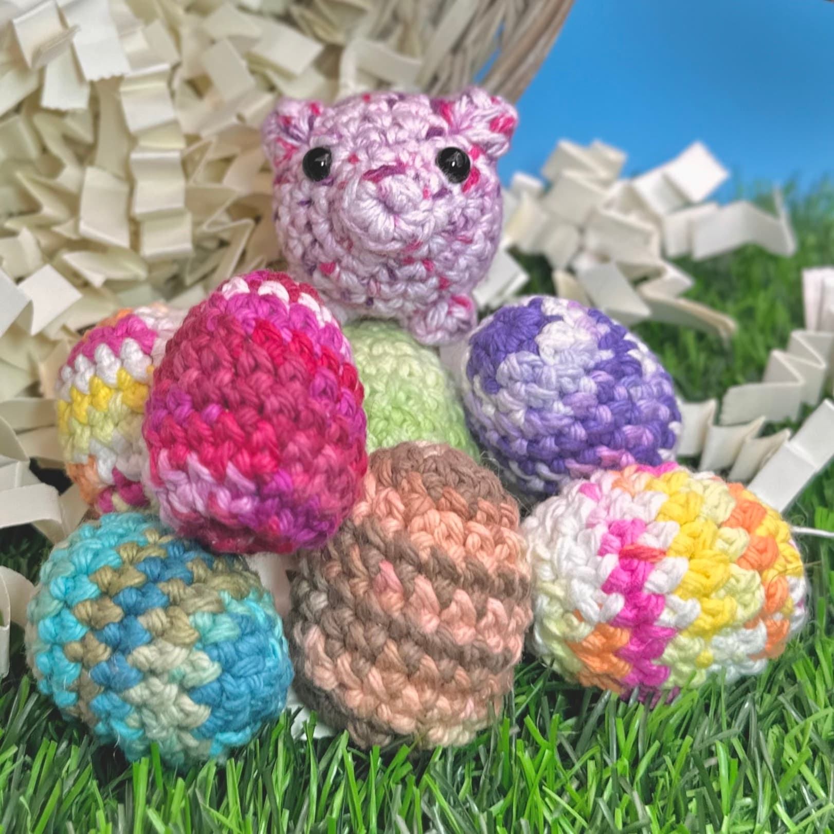 Seven colorful crocheted eggs in pink, purple, blue, brown, and rainbow patterns arranged on artificial grass with a small pink crocheted pig with black bead eyes perched on top.