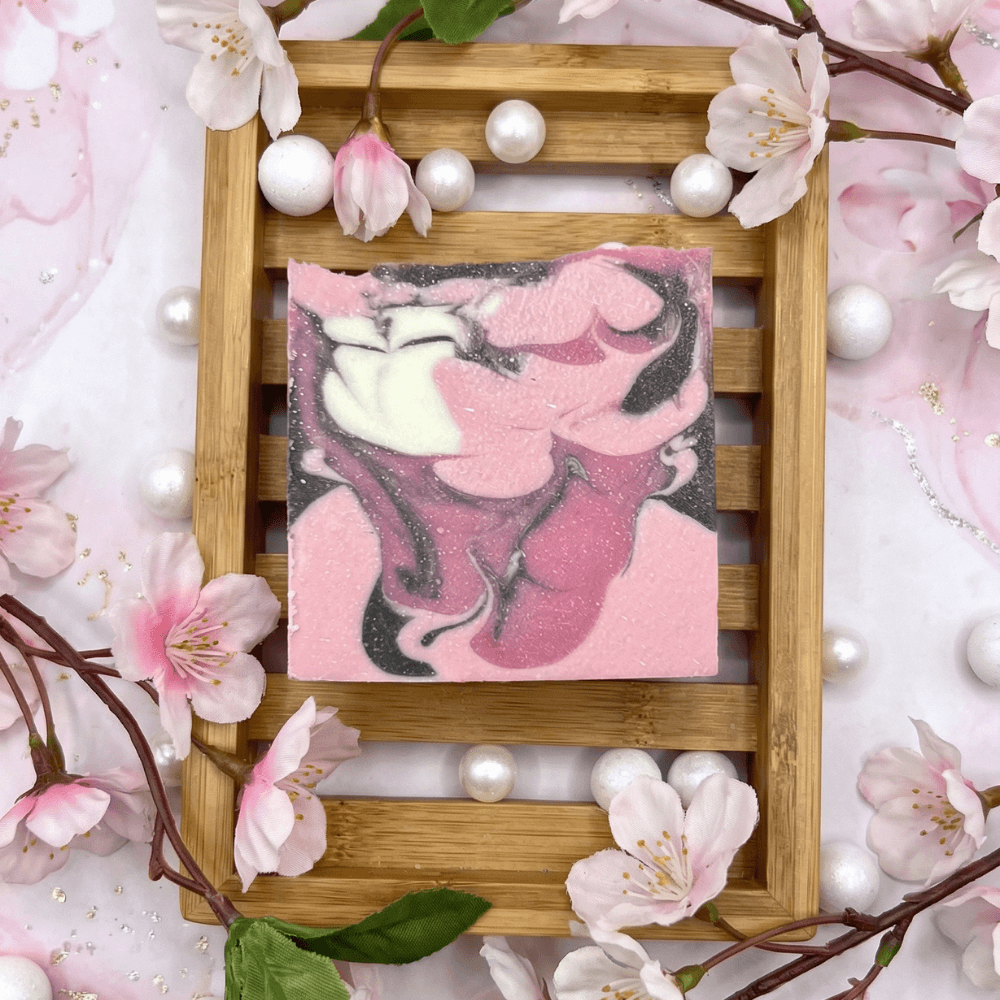 Square handcrafted soap bar with pink, cream, and charcoal swirls resembling cherry blossoms. Displayed on bamboo tray surrounded by white and pink cherry blossom flowers and pearl accents.