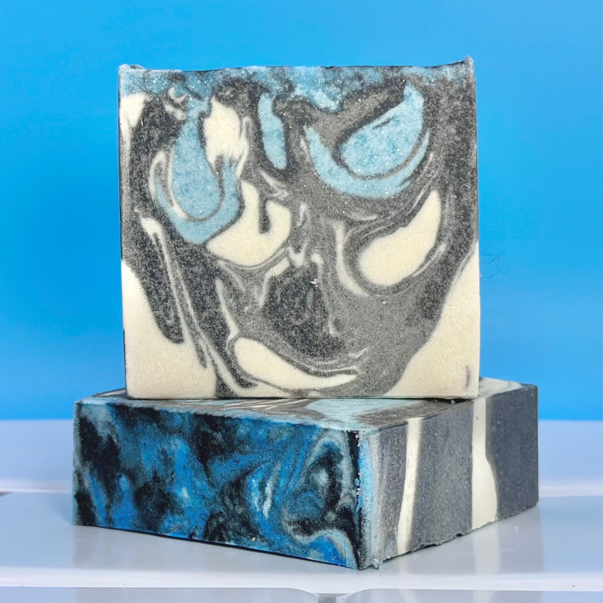 Three bars of handcrafted soap with swirled patterns in charcoal gray, white, and bright blue, stacked against a blue background. The top bar shows marbled texture with activated charcoal.