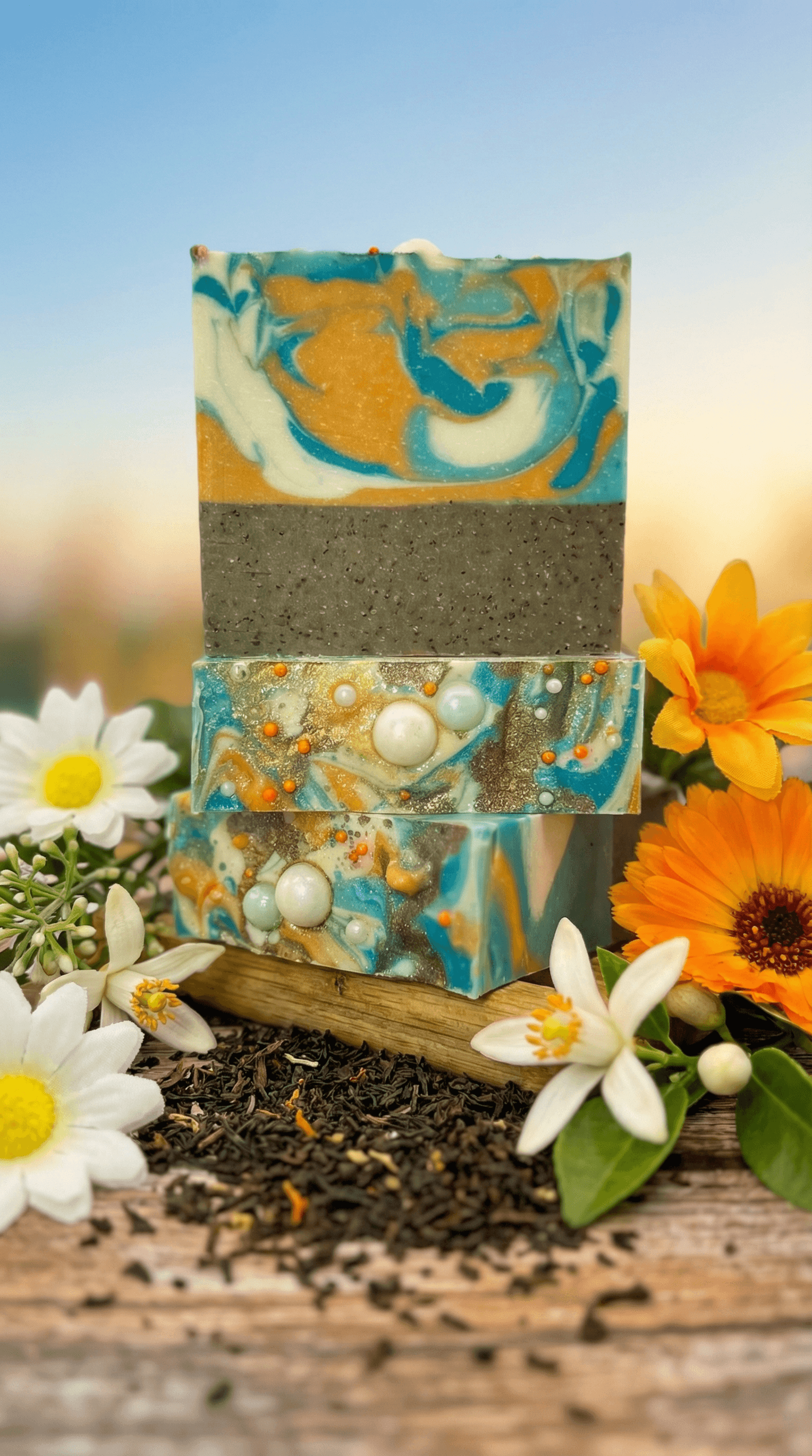 Three stacked handmade soap bars with swirled turquoise and orange layers on top, dark grey speckled middle layer. Bottom bars feature embedded pearl beads and orange dots. Surrounded by white and yellow flowers and loose tea leaves.