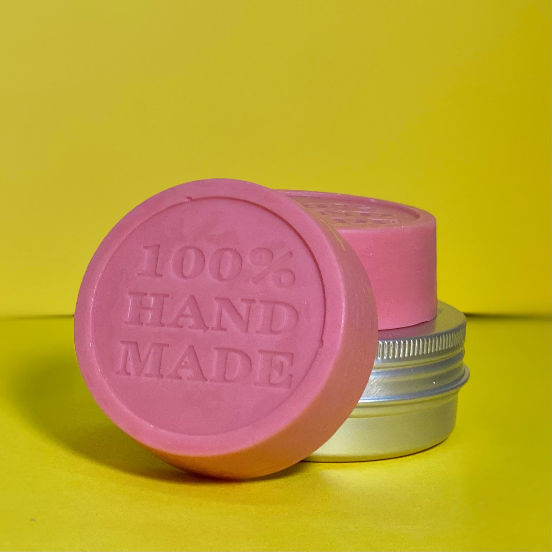 Pink circular lotion bar with embossed text reading '100% HAND MADE' standing upright against yellow background, with two additional bars and small tin visible behind it.