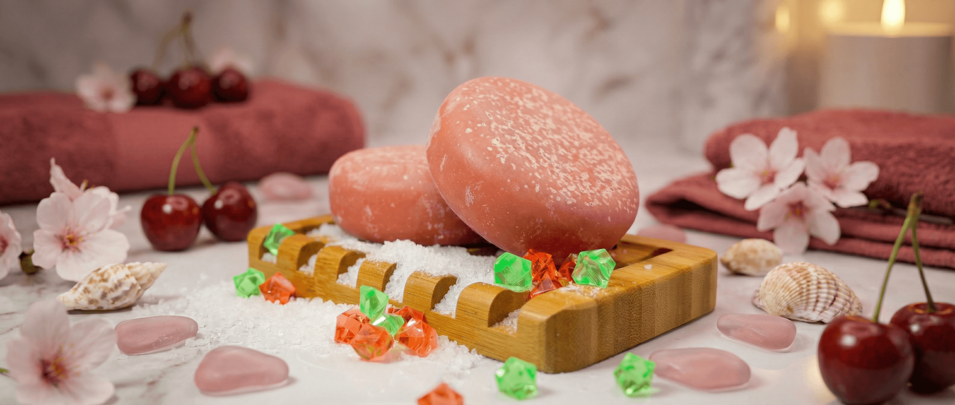Pink heart-shaped sea salt scrub bar with glistening crystals resting on bamboo soap dish, surrounded by fresh cherries, cherry blossoms, seashells, and colorful decorative gems.