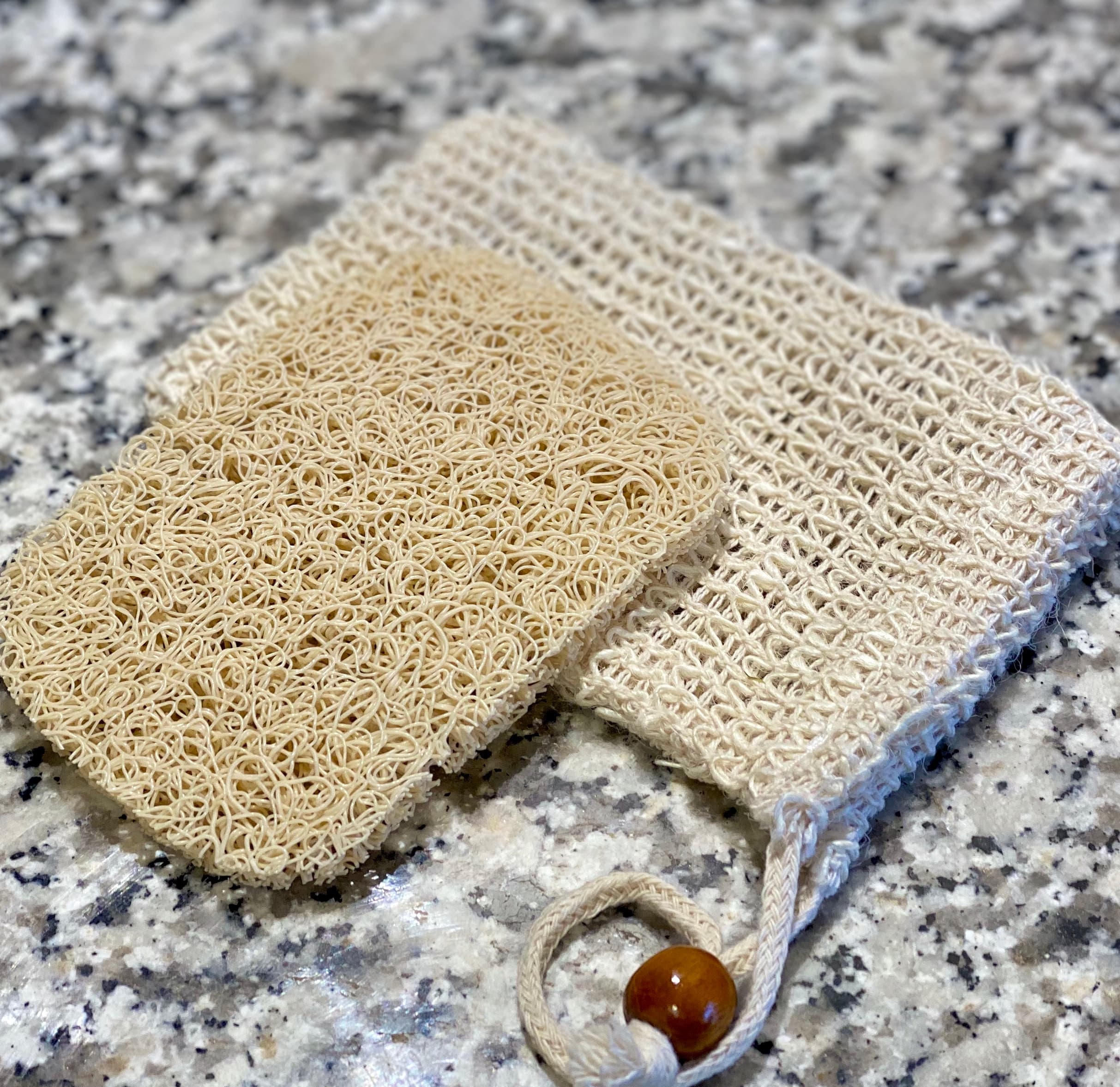 Two natural fiber cleaning tools on granite: a looped sisal scrubber with textured loops and a flat mesh soap saver bag with wooden bead closure in cream tones.