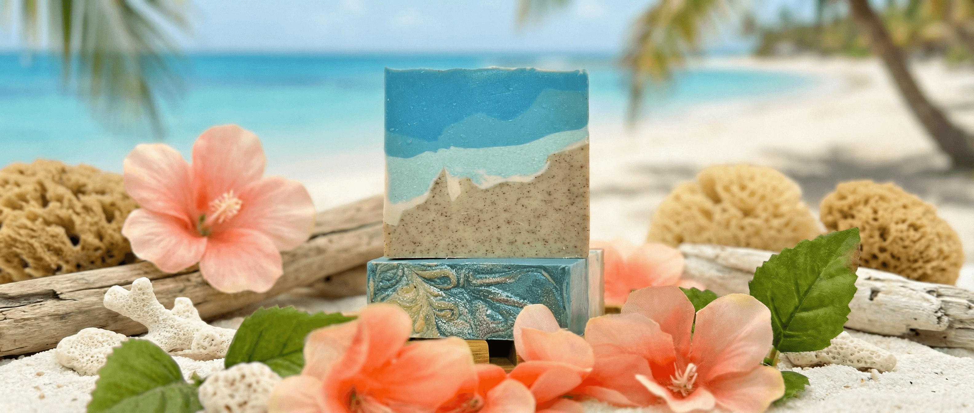 Handcrafted soap bar with layered ocean-blue top and sandy beige bottom with visible exfoliating specks, displayed on driftwood with coral hibiscus flowers, sea sponges, and beach backdrop.